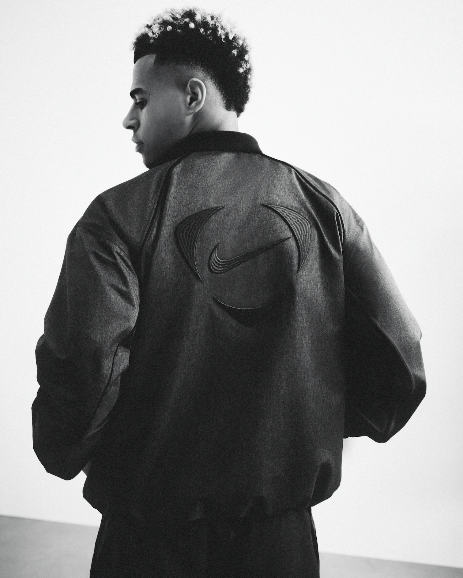 Black and white photo of a man with short curly hair wearing a Nike jacket with logo on the back, facing left with his head slightly tilted downward.