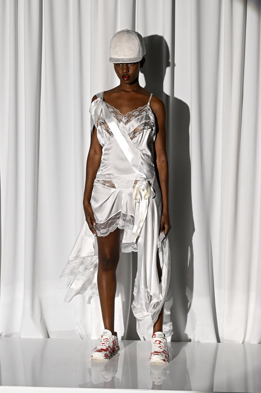 Model wearing white satin lingerie with lace details, paired with a white baseball cap, white sneakers, in front of a white curtain backdrop.