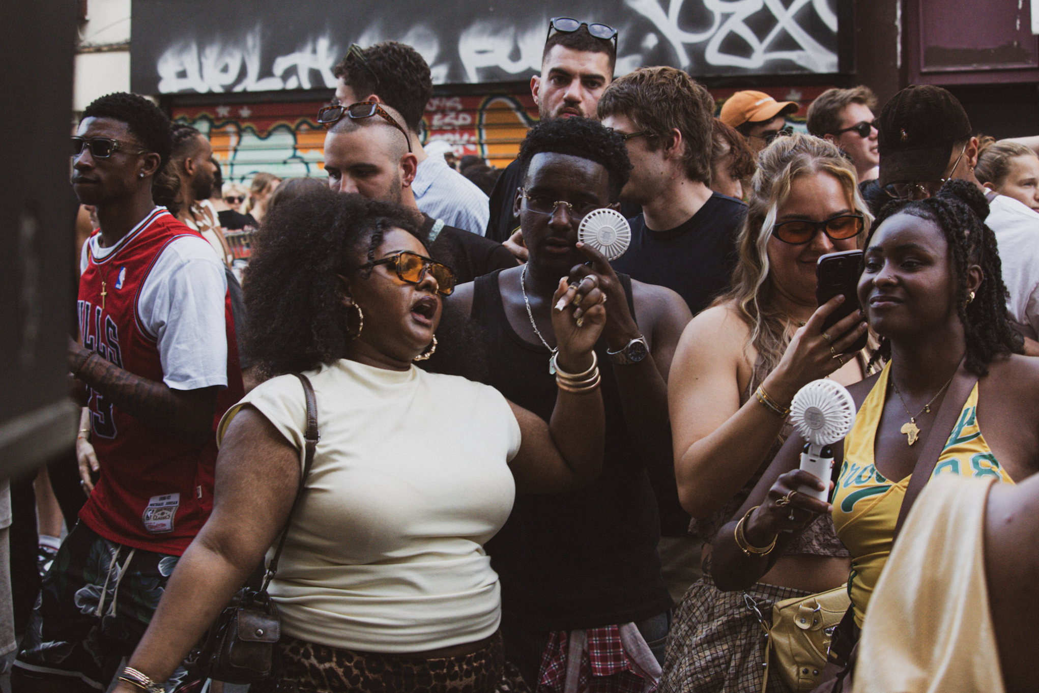 A diverse crowd of people gathered outdoors, some wearing sunglasses, with a brick wall featuring graffiti in the background.