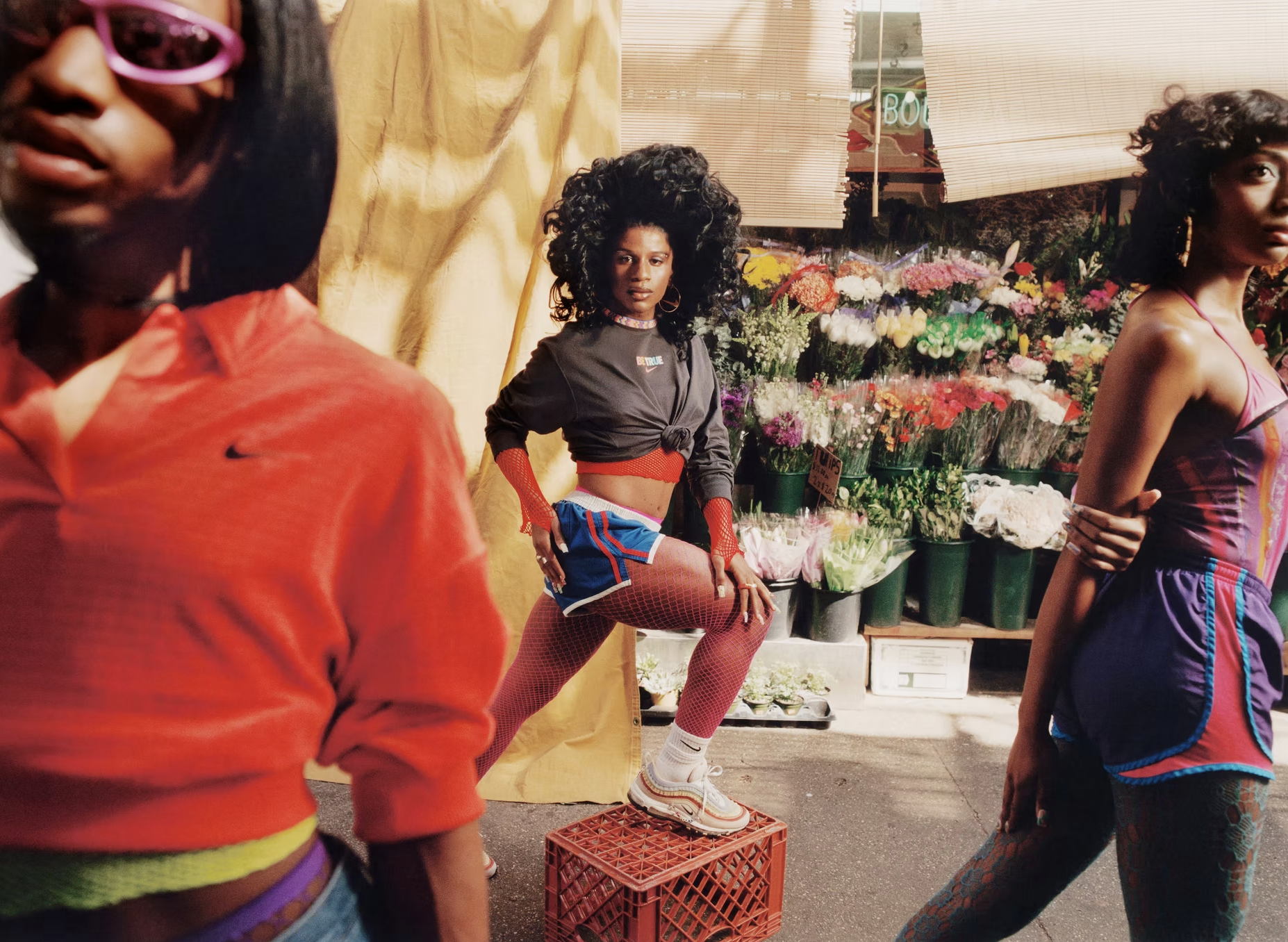 A woman with voluminous curly hair, wearing a sporty outfit with shorts, fishnet stockings, a black jacket, and sneakers, poses with one leg resting on a red crate inside a flower shop. Other women are partially visible around her, and a variety of colorful flowers in pots are displayed in the background.