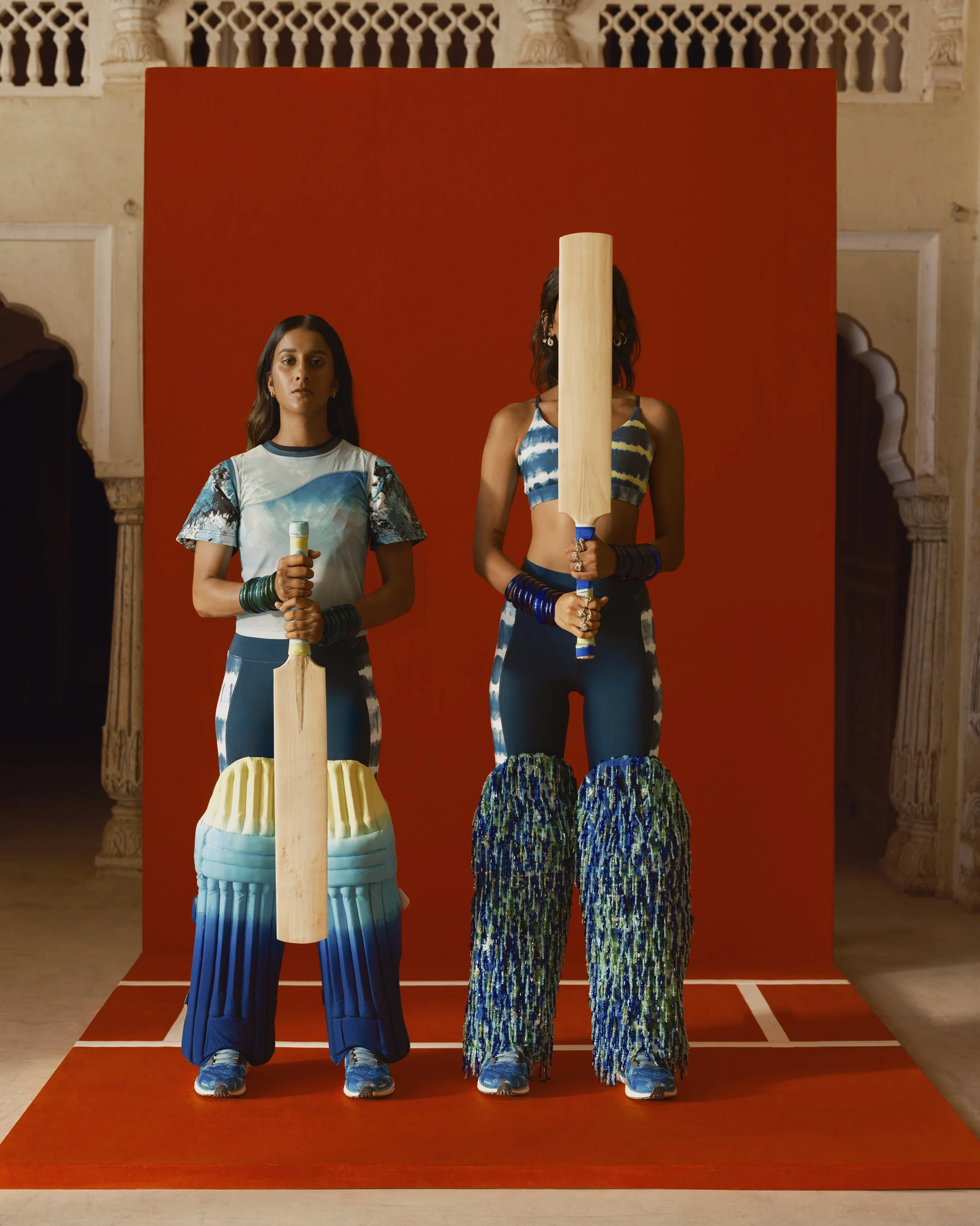 Three women standing behind a red backdrop, holding wooden paddles, wearing colorful and patterned clothing, and surrounded by traditional architectural elements.
