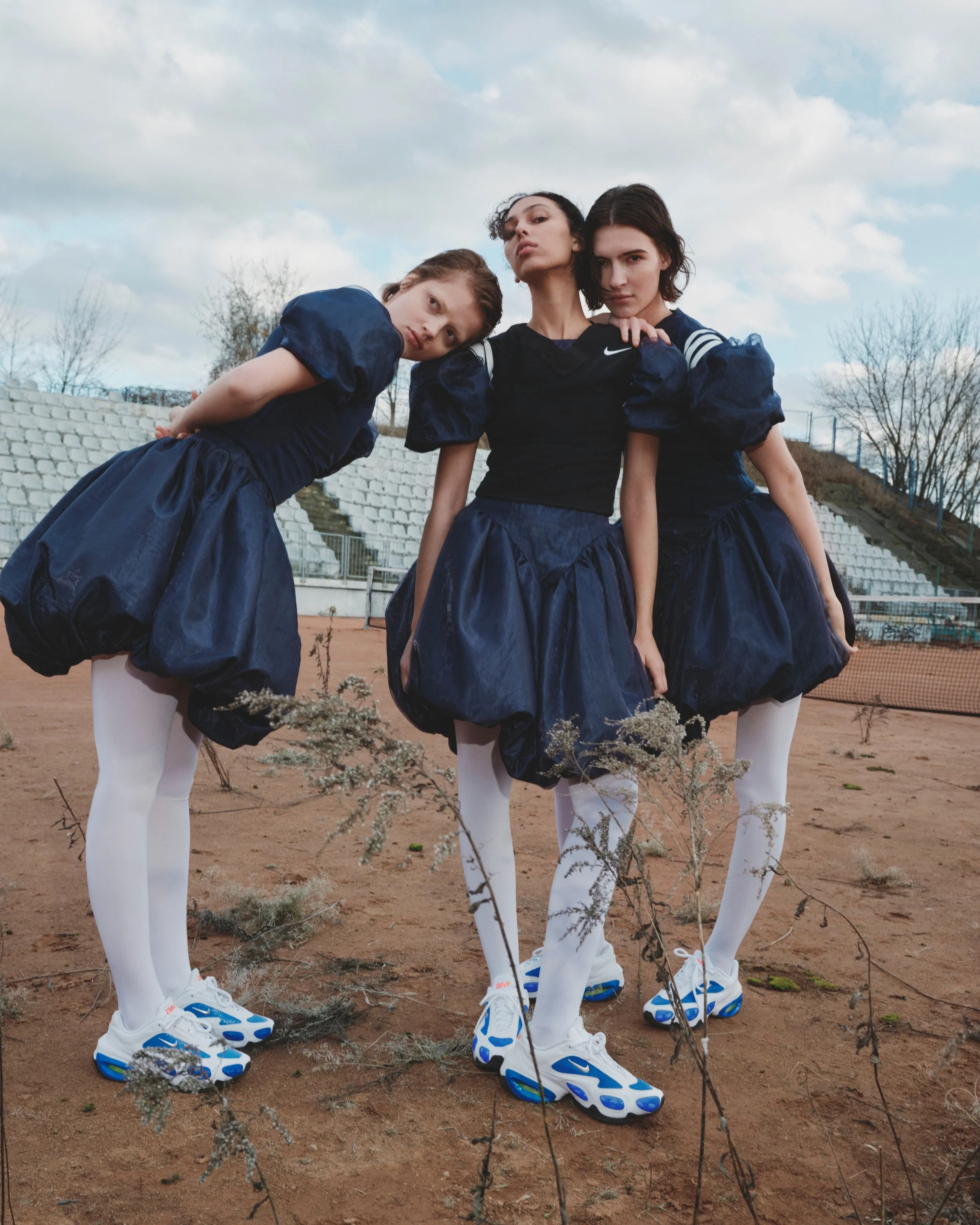 Three young women in dark sports uniforms and white tights, wearing white and blue sneakers, posing outdoors on a dirt field with dry plants in the foreground and empty stadium seats in the background.
