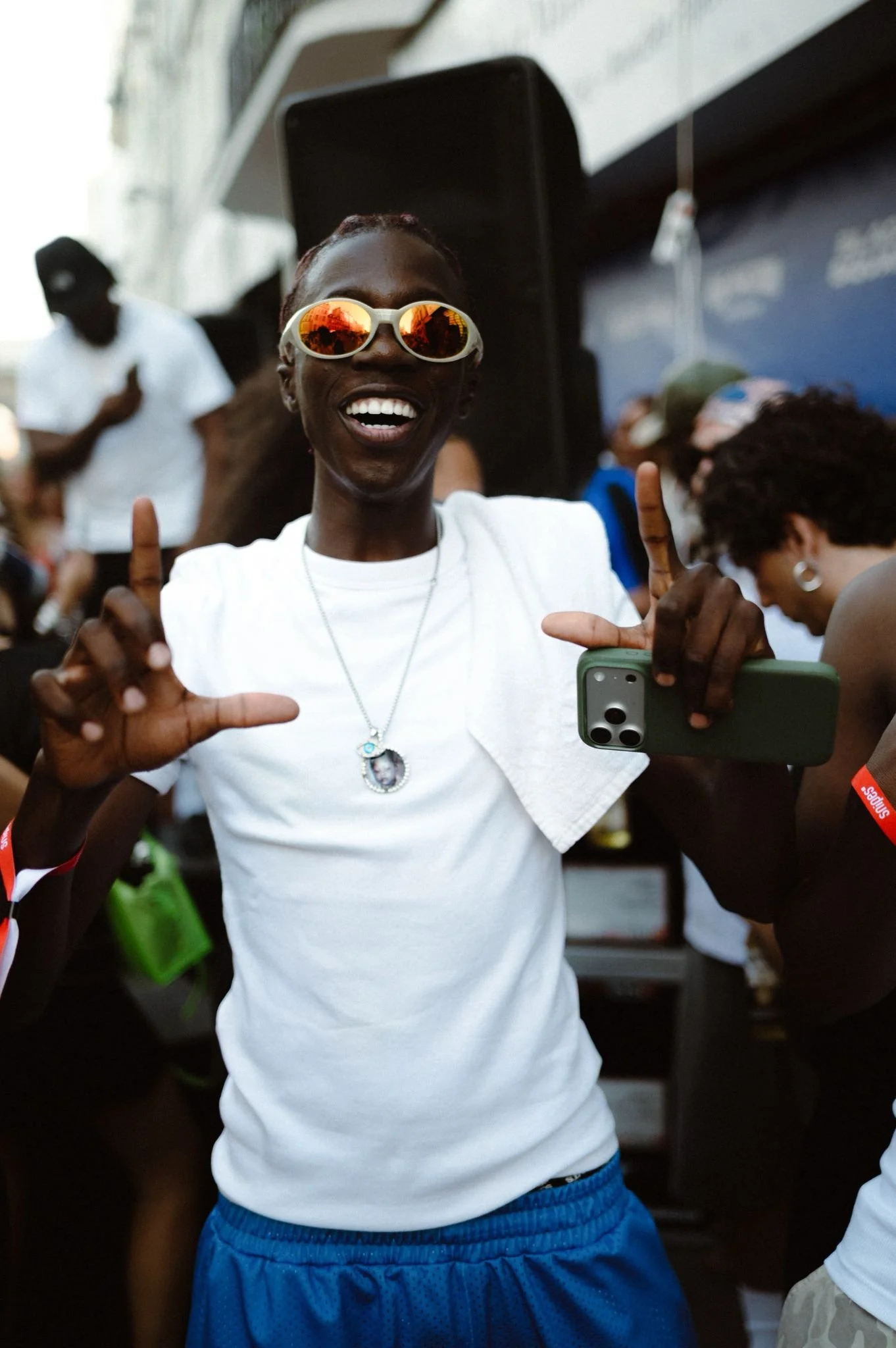 A person smiling widely, wearing sunglasses with red reflective lenses, a white t-shirt, and blue shorts, holding a green phone, and making a hand gesture with both hands, outdoors at a social event.
