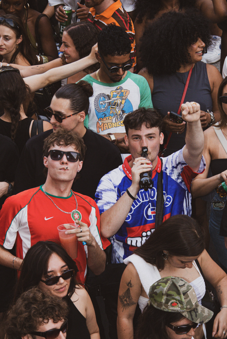 A crowd of diverse young adults at an outdoor event or concert, some holding drinks and wearing casual clothing with sports jerseys and accessories, enjoying the lively atmosphere.