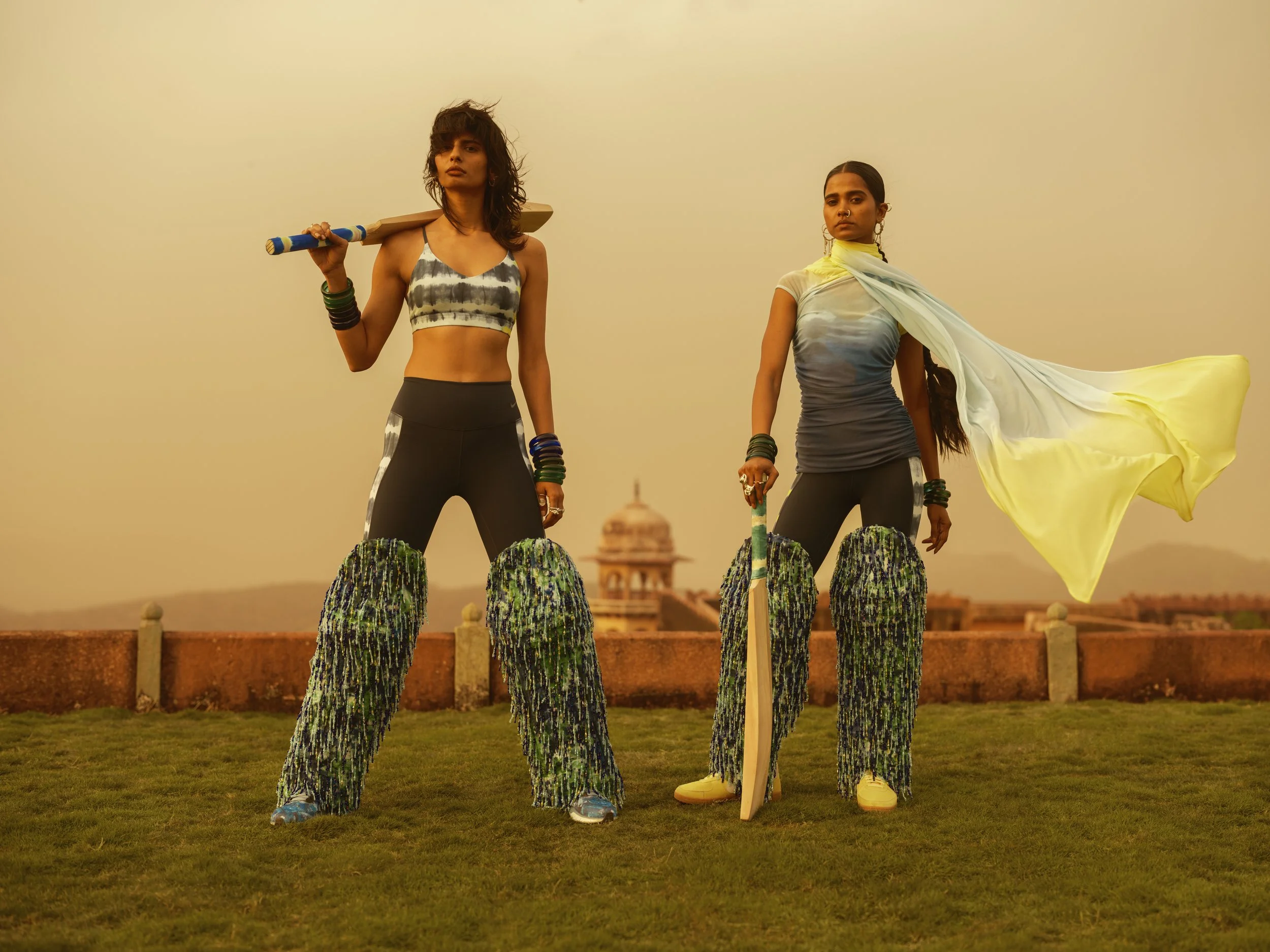 Two women dressed in sporty attire standing on a grassy area during sunset, each on decorated stilts, with one holding a flag and the other with a skateboard. A historic monument is visible in the background.