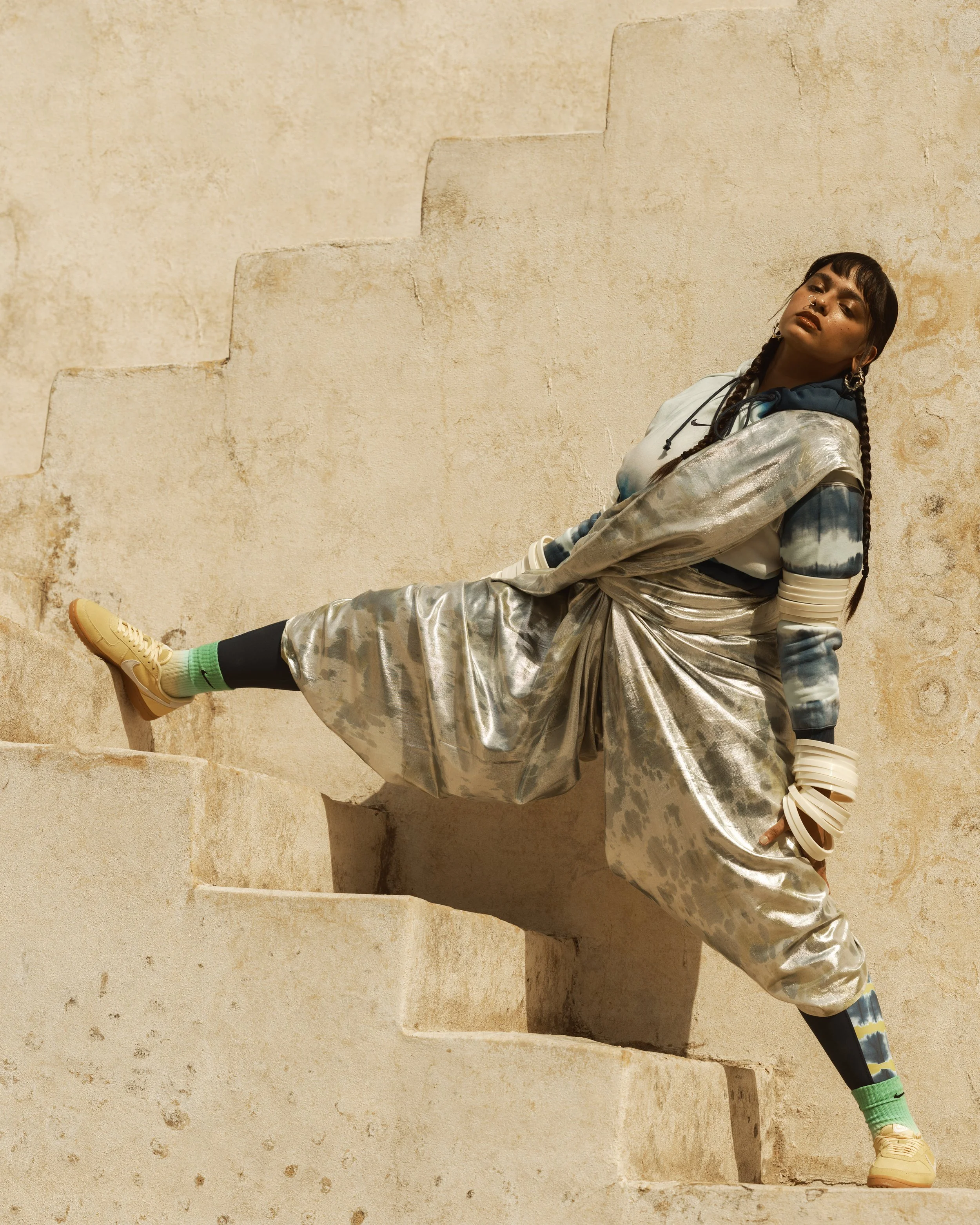 A woman leaning against a beige textured wall with stairs in the background, wearing a shiny silver jacket, patterned pants, and beige shoes.