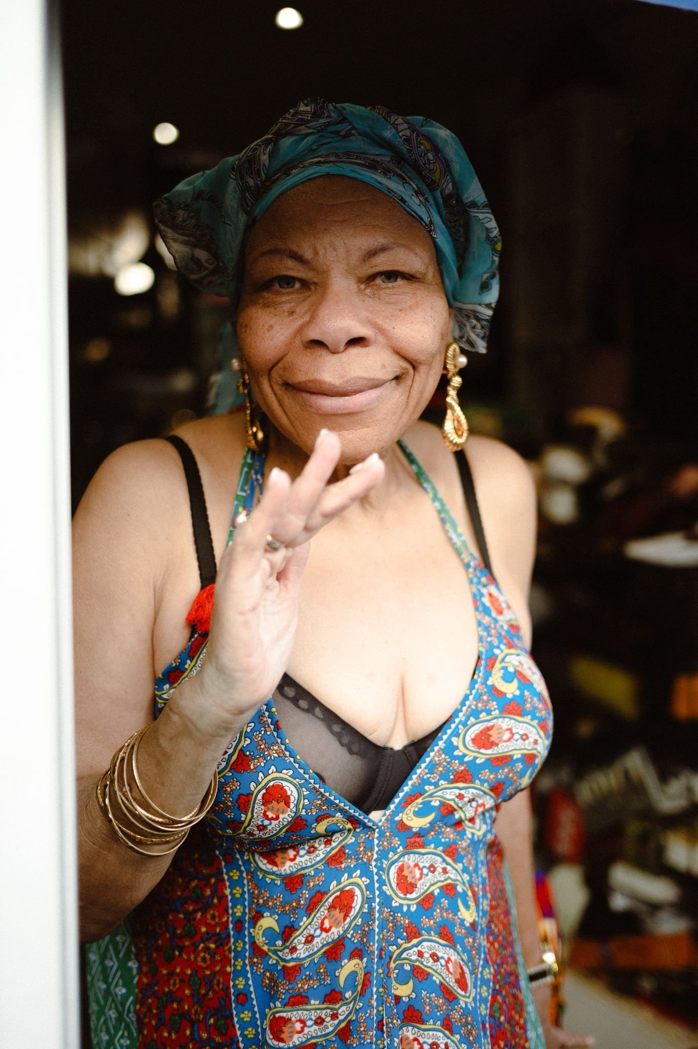 Close-up of a smiling woman wearing a blue patterned dress and headscarf, with jewelry, standing indoors.