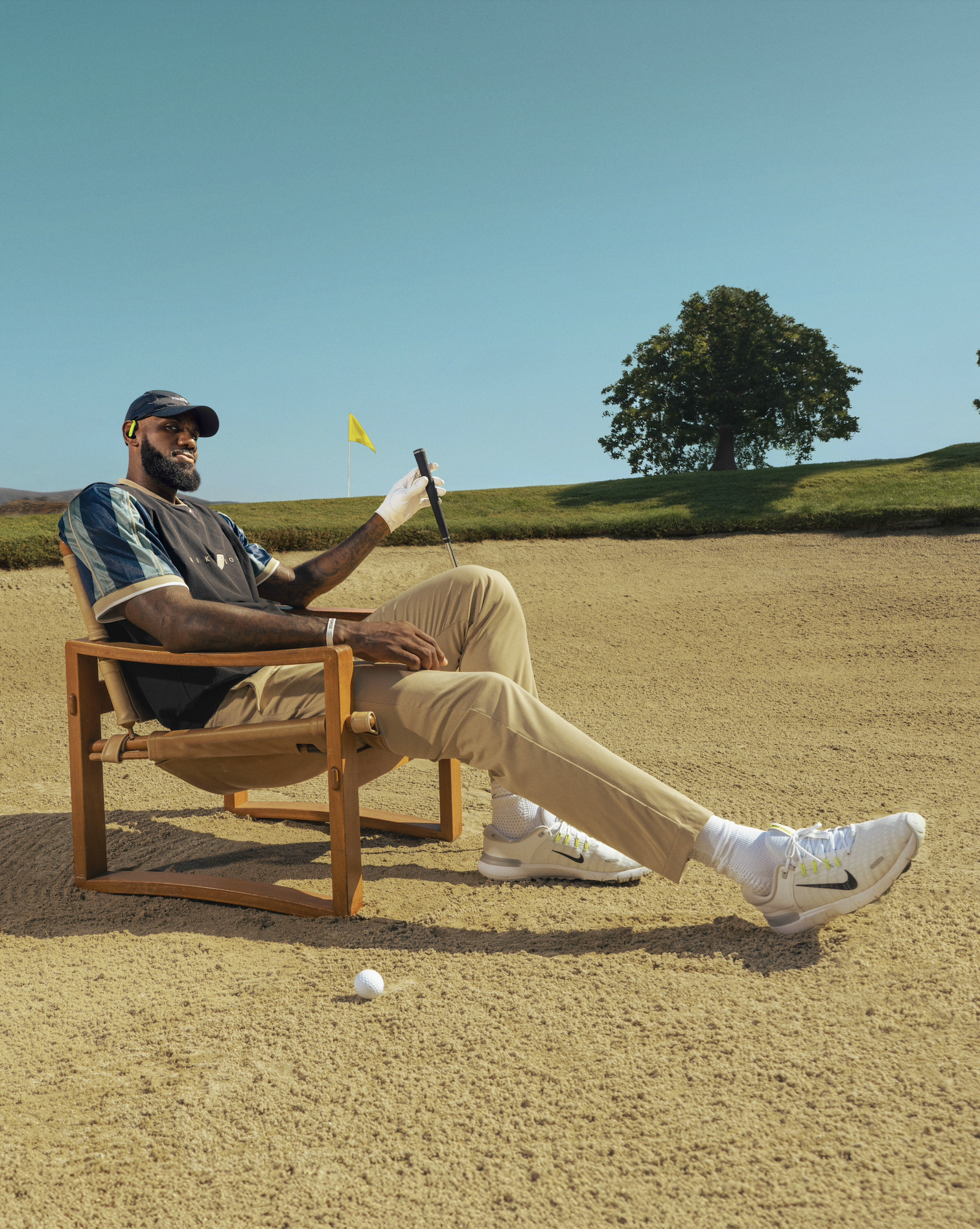 A man sitting on a patio chair on a golf course preparing to putt a golf ball in a sand trap with a yellow flag in the background and a tree on the grassy hill behind him.