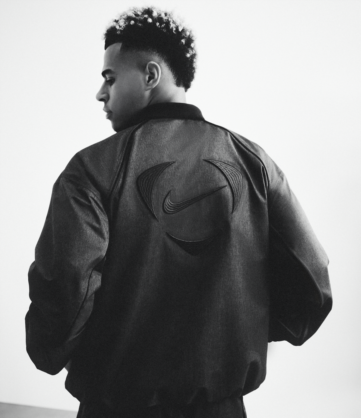Black and white photo of a man facing left, wearing a black jacket with Nike logos on the back, against a plain white background.