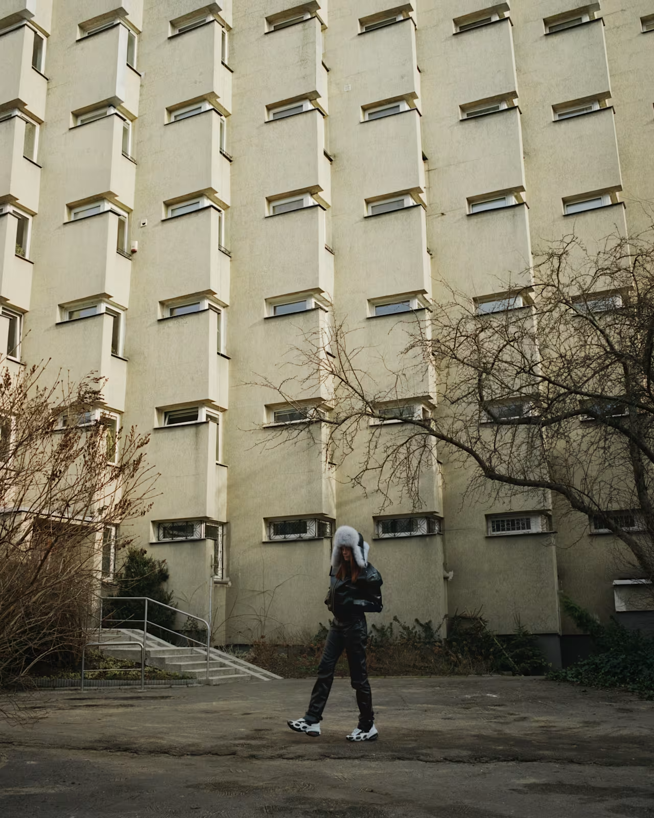 A person wearing a furry hat riding rollerblades on a paved area in front of a beige apartment building with small windows and leafless trees.