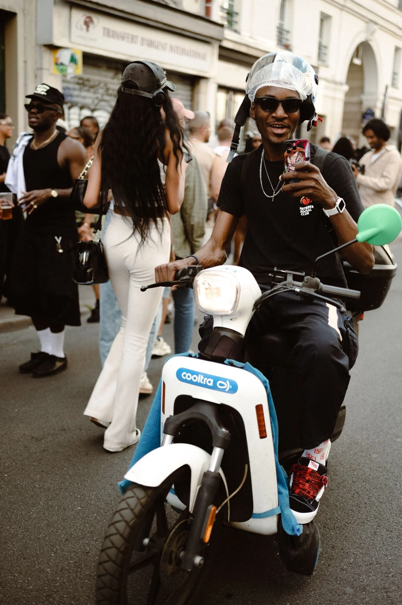 A man wearing a helmet, sunglasses, and a black t-shirt with a logo, sitting on a small white and blue electric scooter, smiling at the camera, in a busy city street.