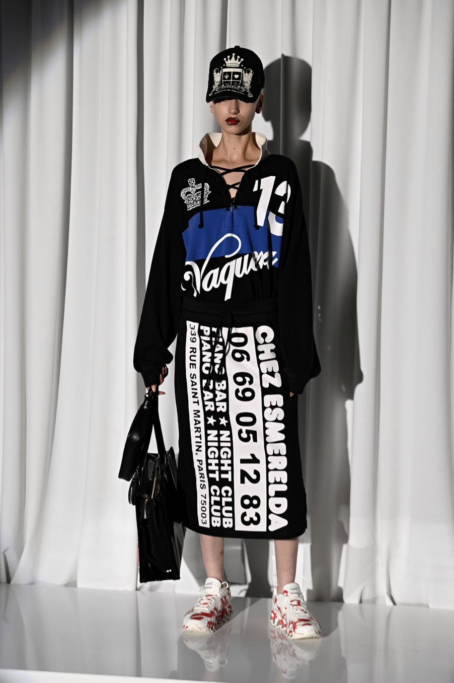 Model wearing black, blue, and white streetwear outfit with printed text and graphics, including a cap, hoodie, and long skirt, standing on a glossy white floor against a backdrop of white curtains.