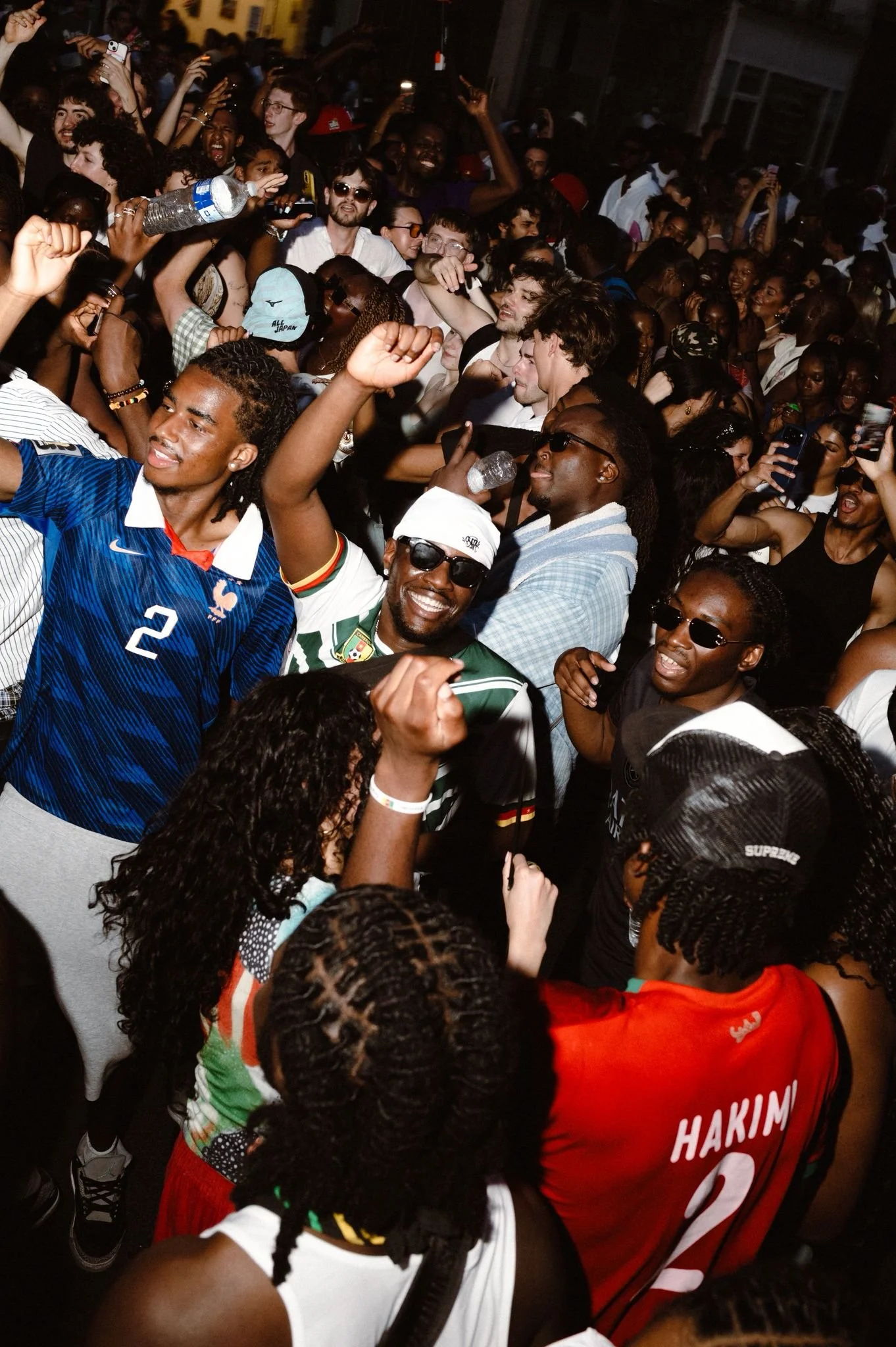 A large crowd of people gathered at night, many smiling and taking photos, with some holding water bottles. The scene appears lively and energetic.