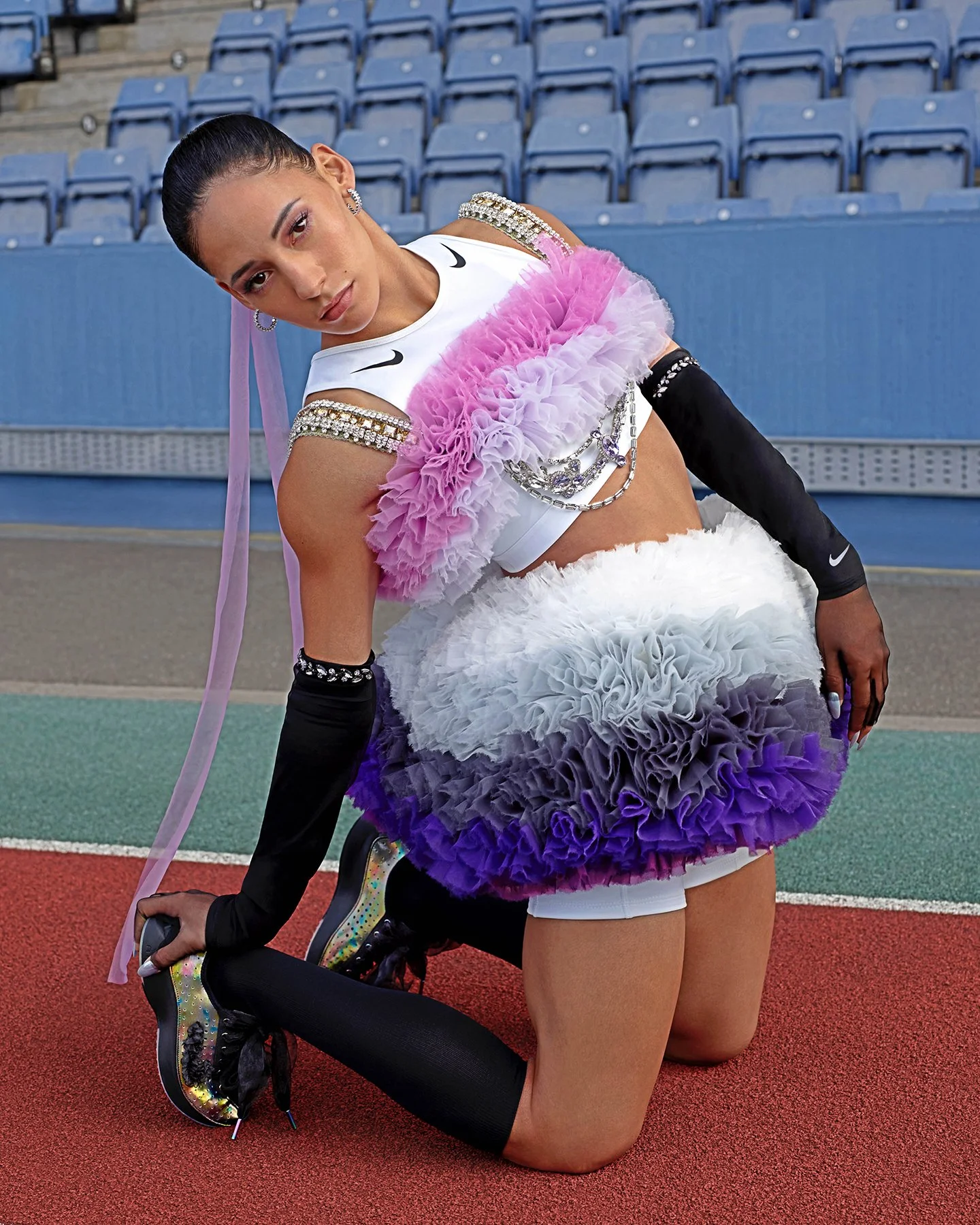 A female athlete dressed in a colorful, ruffled skirt and athletic gear, kneeling on a track field.