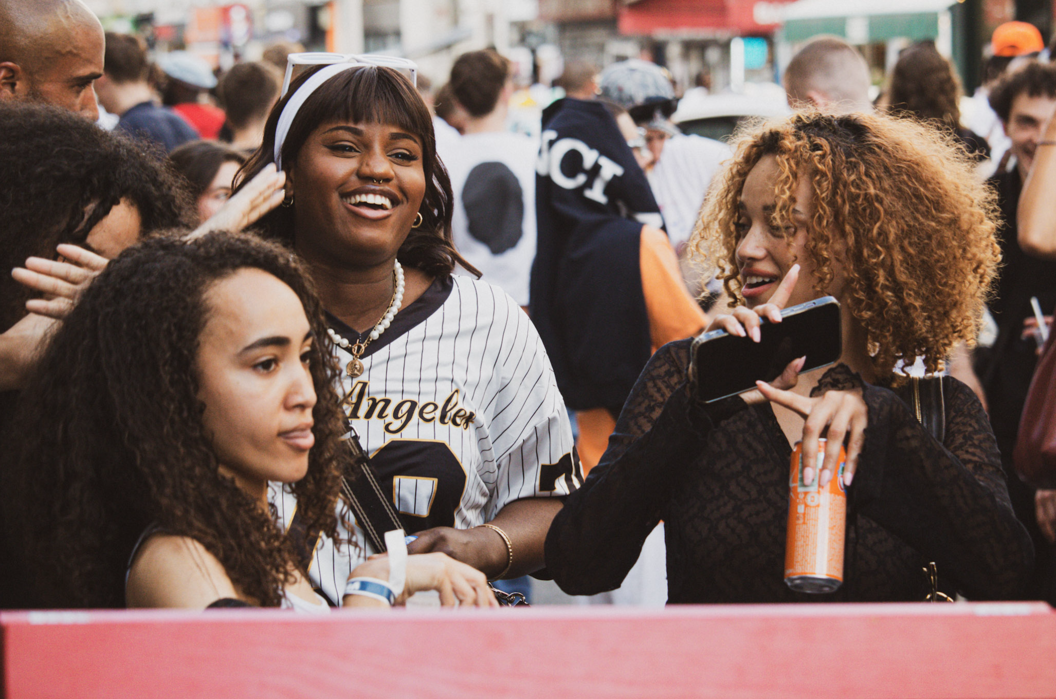 A group of diverse people, including women and children, gathered outdoors during daylight. One woman is wearing a baseball jersey with 'Angeles' on it, smiling and wearing sunglasses on her head. Another woman with curly red hair and tattooed arms is holding a smartphone and a soda can. The scene appears lively, with many people in the background, some holding umbrellas, in an urban setting.
