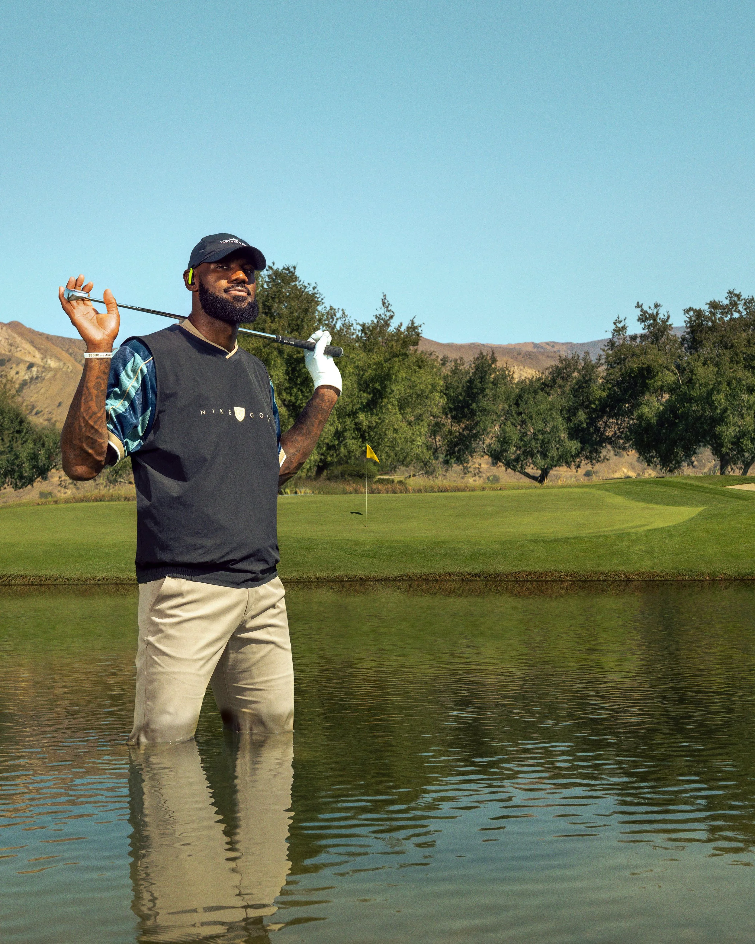 A man standing knee-deep in water on a golf course, holding a golf club over his shoulder, with trees, hills, and a clear blue sky in the background.