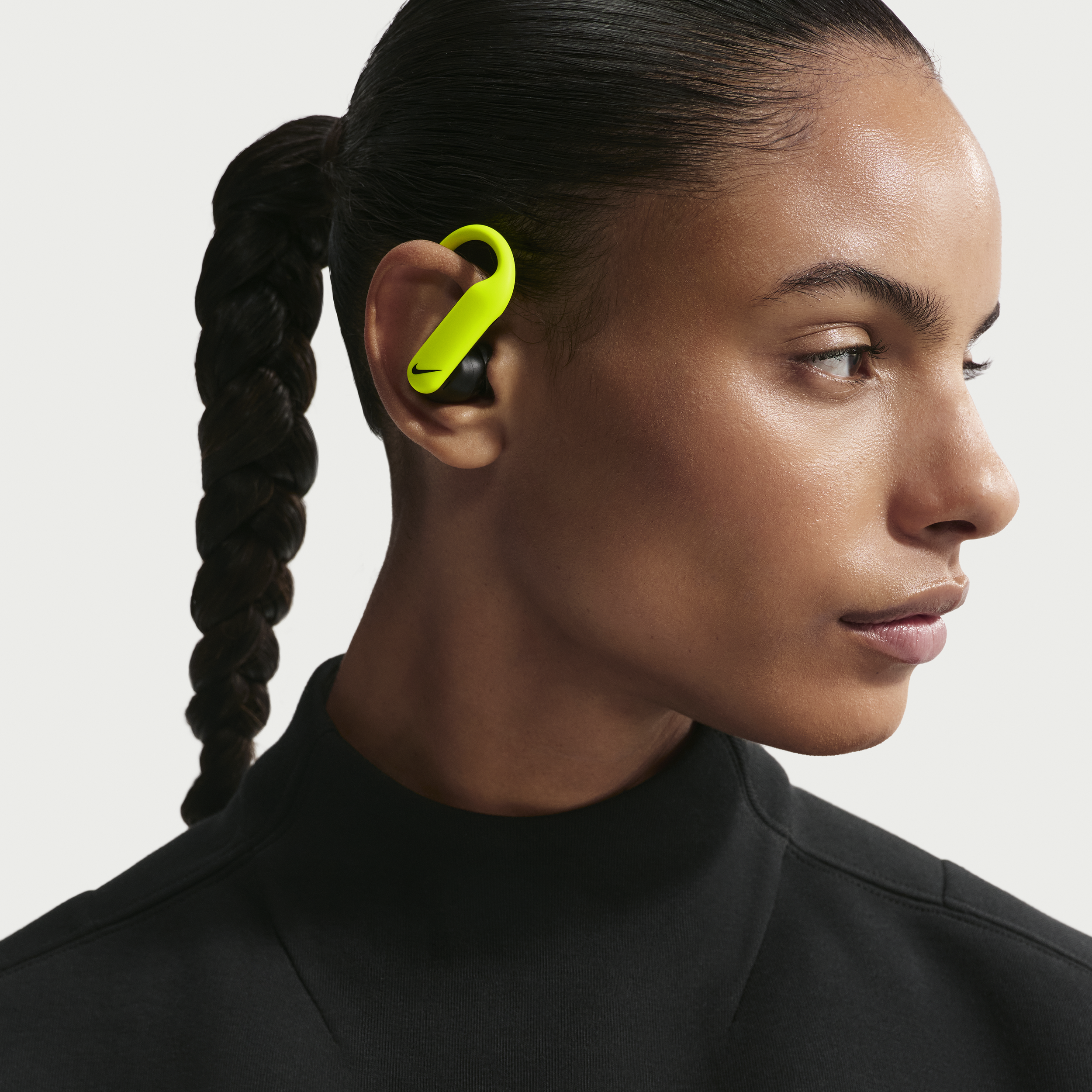 Close-up of a woman with dark hair tied in a braid, wearing a neon green Nike wireless earbud in her right ear, dressed in a black top.
