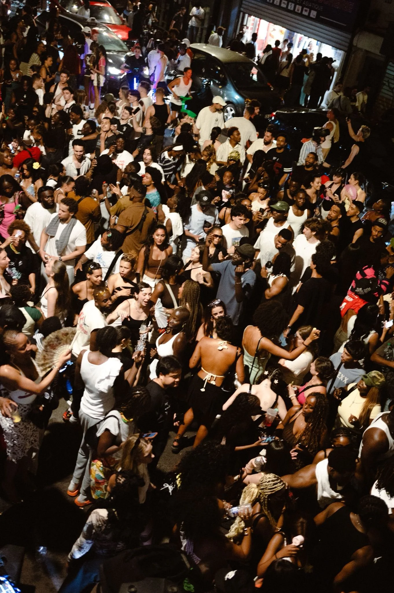 Crowd of people gathered on a lively city street at night, some looking at their phones, standing and walking around near parked cars and shop entrances.