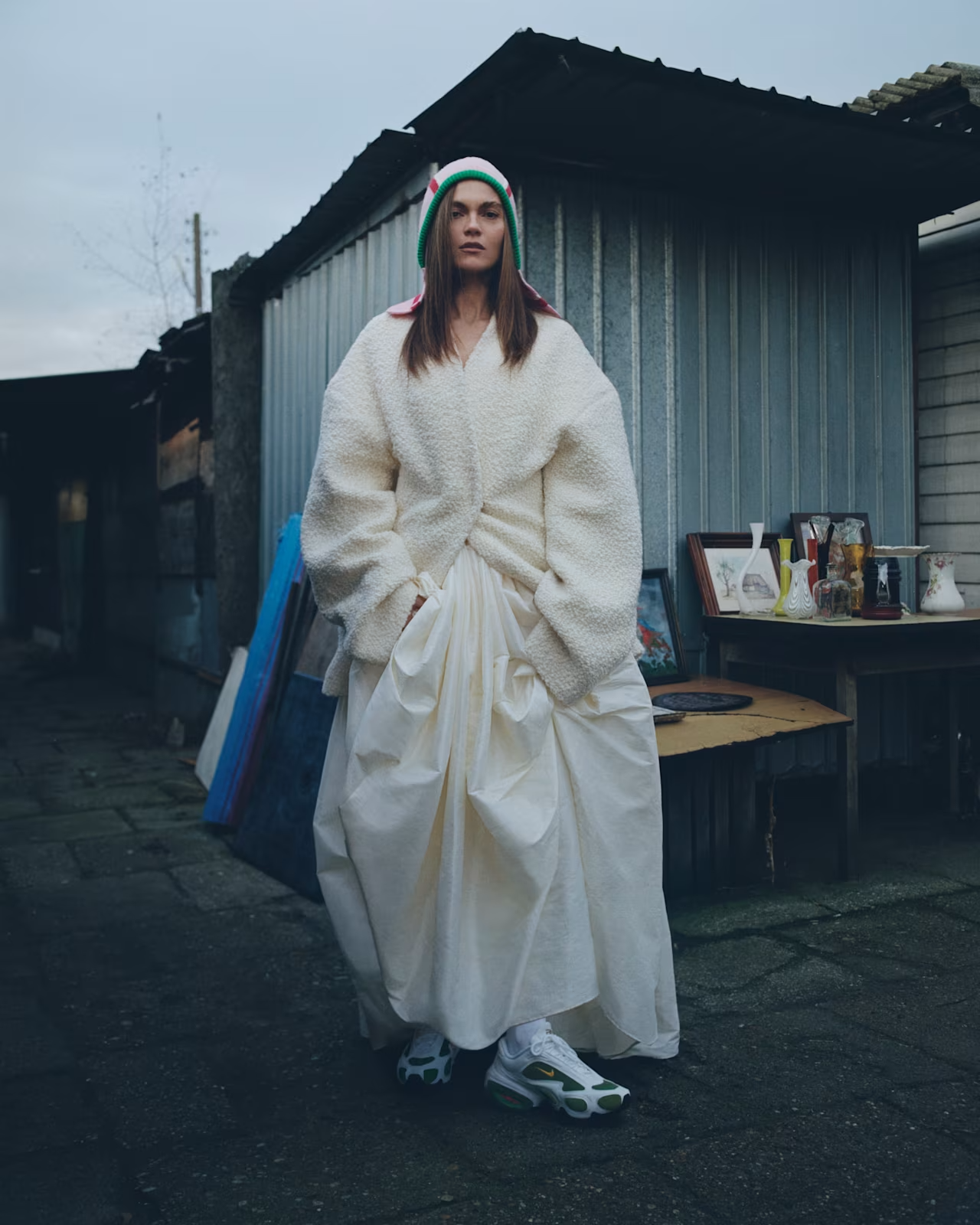 A woman standing outside in a yard, wearing a green, white, and red knit beanie, a white fuzzy jacket, and a long flowing white skirt, with sneakers. There is a table behind her with various decorative items and framed pictures.