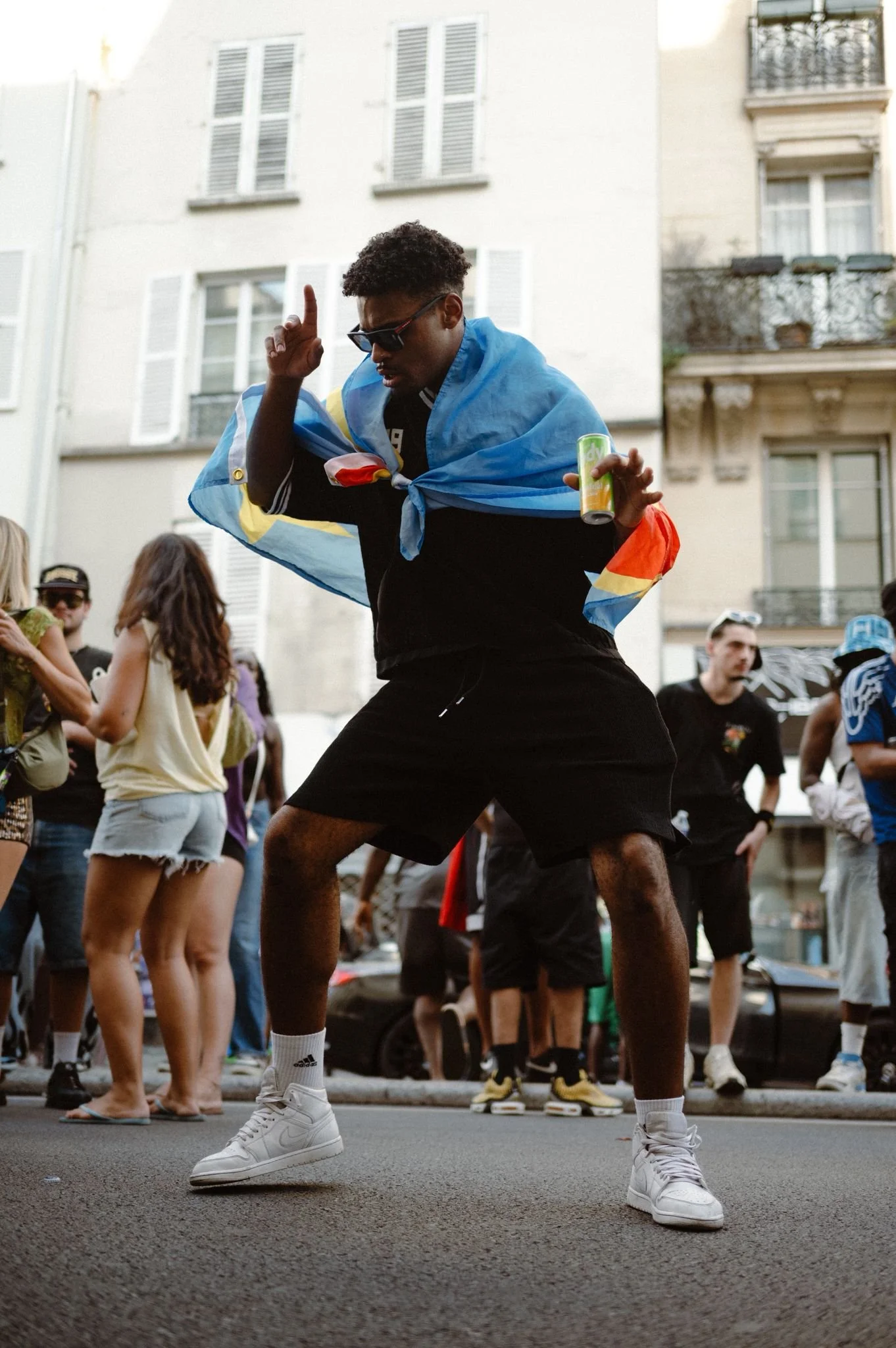 A person dancing in front of a crowd, wearing a colorful flag as a cape, sunglasses, black shorts, white sneakers, and holding a drink, with other people in the background on a city street.