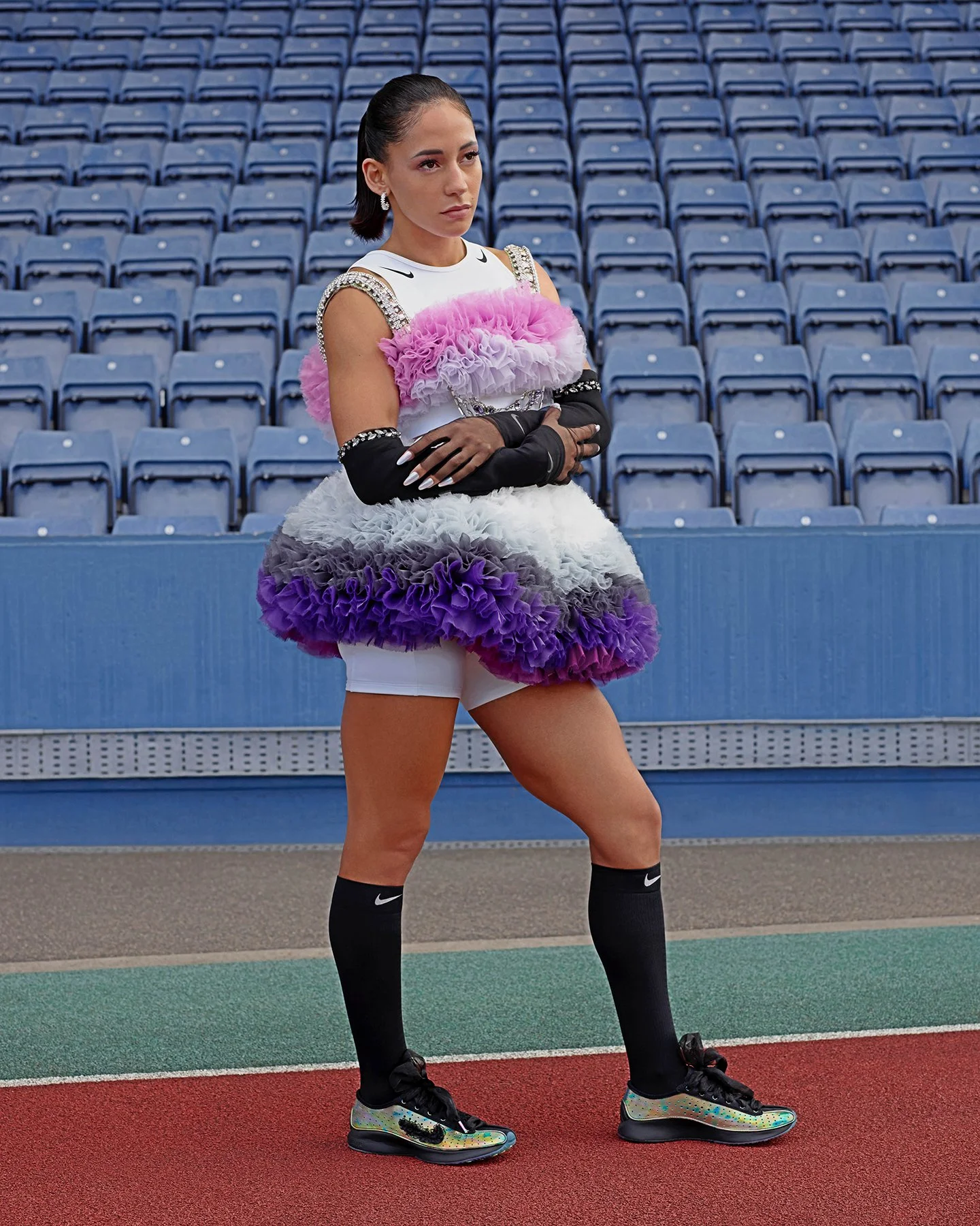 Female athlete standing on a track at a stadium with empty seats, wearing a colorful, ruffled sports dress with black gloves, knee-high socks, and sports shoes.