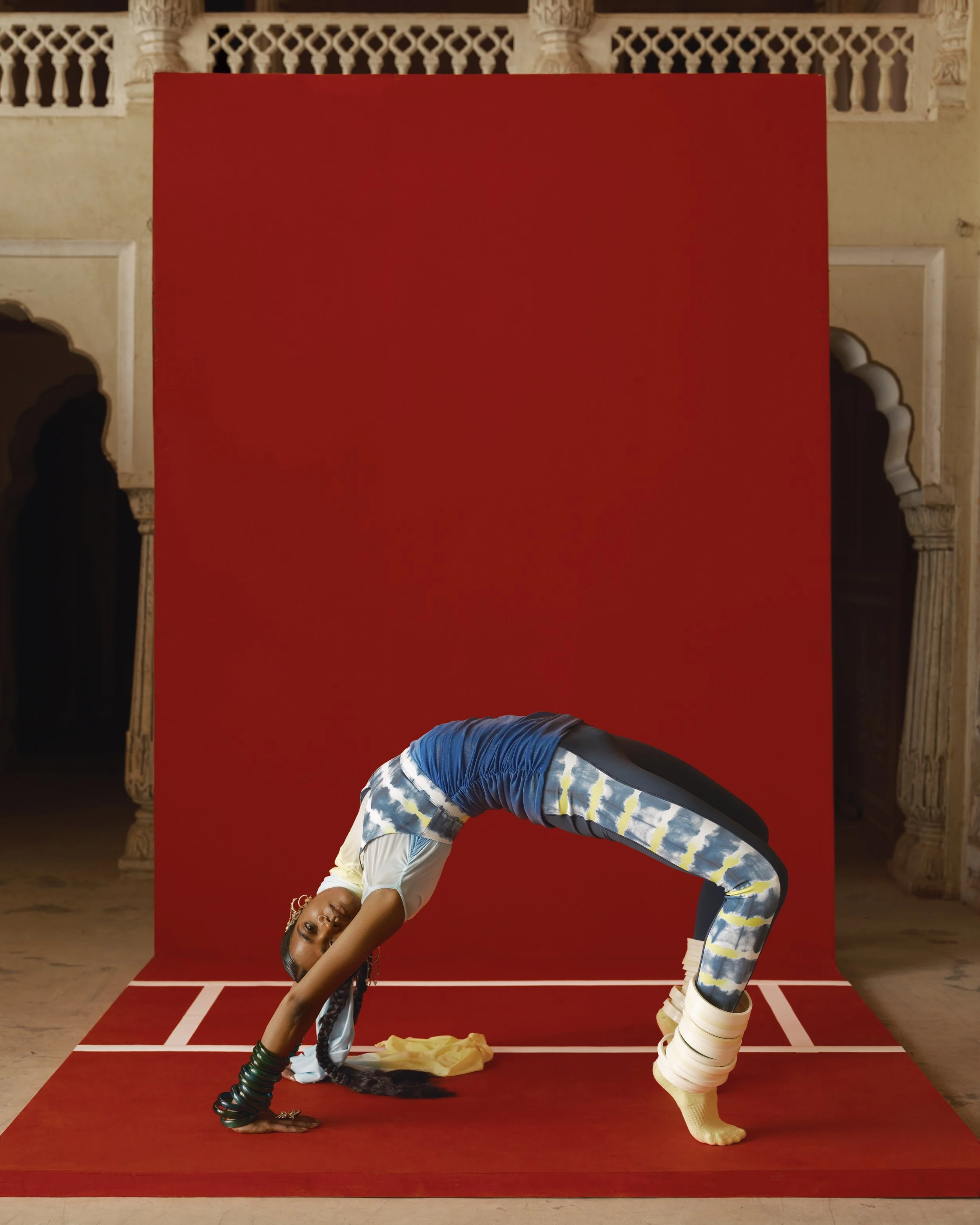 A woman performing a yoga pose on a red yoga mat in front of a red backdrop. She is bent forward with her hands on the ground and one leg raised and bent at the knee. She is wearing colorful athletic clothing and white shoes, with her hair in braids.