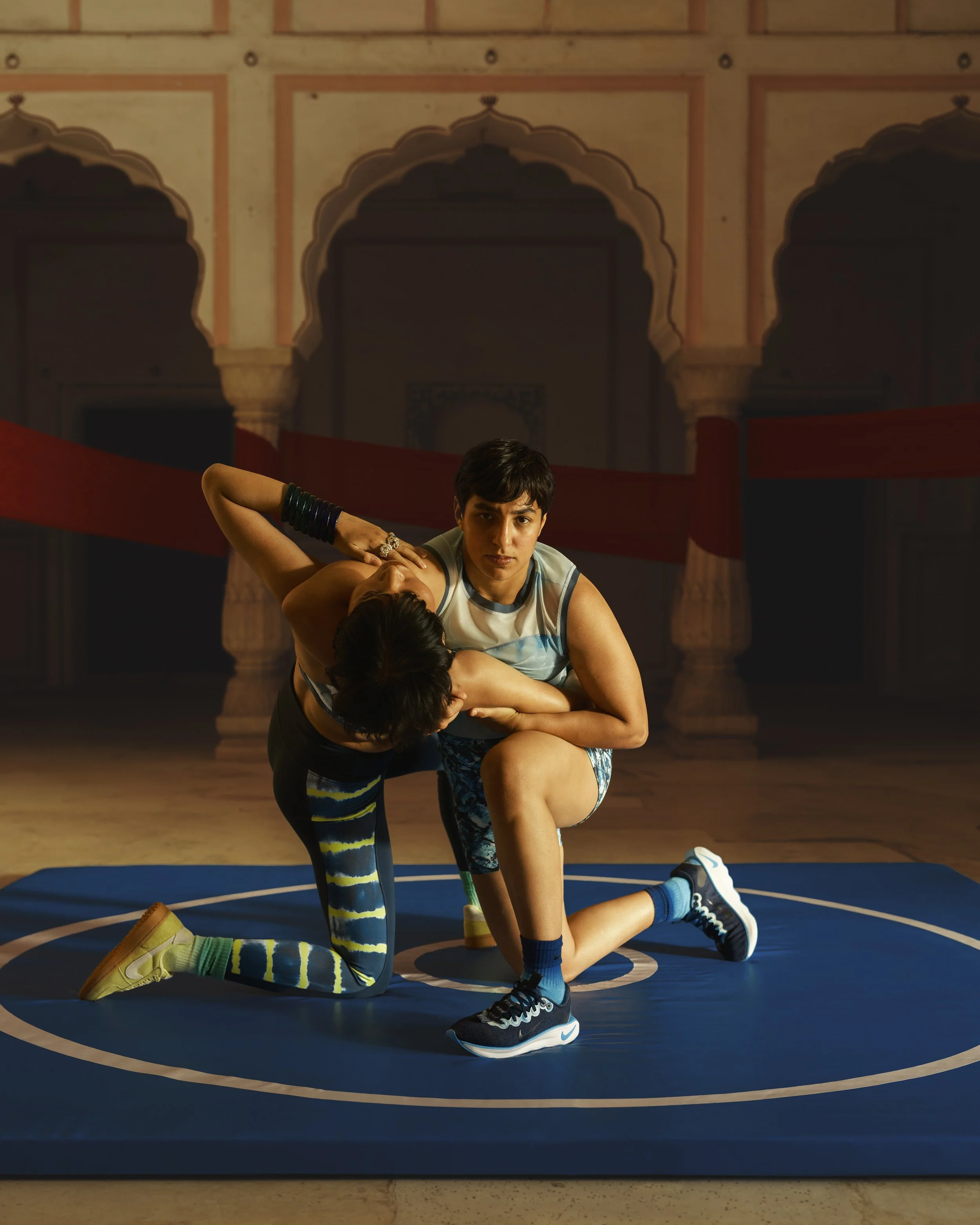A young person with short dark hair practicing wrestling or grappling on a blue wrestling mat. They are wearing striped leggings, a sleeveless top, and sneakers. The background features traditional Indian or Mughal architecture with ornate arches and pillars.