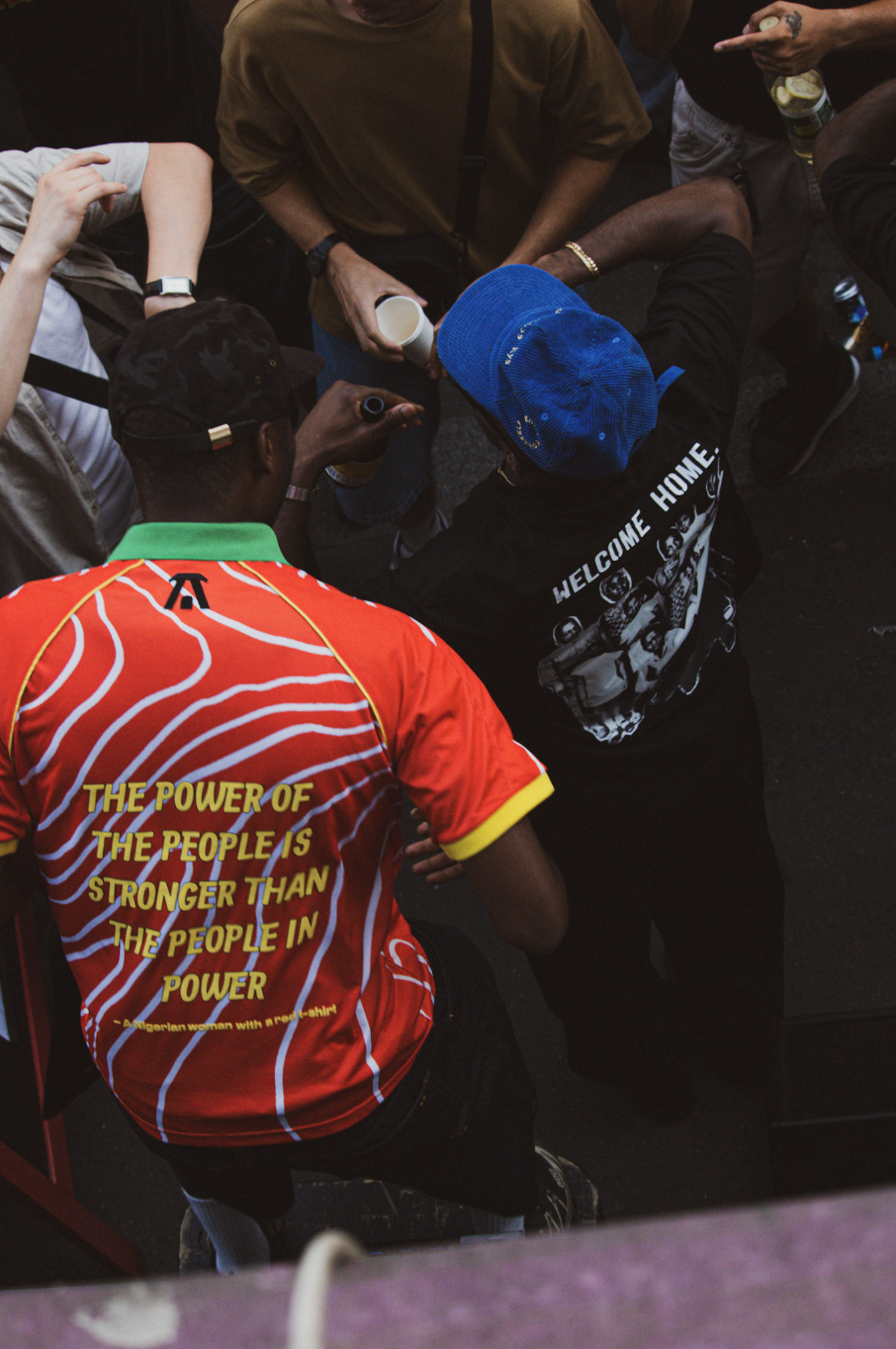 A group of people gathered together, with some holding drinks. One person is wearing a red and orange shirt with the quote, "The power of the people is stronger than the people in power," and another person is wearing a blue cap and a black shirt with "Welcome Home" written on it.