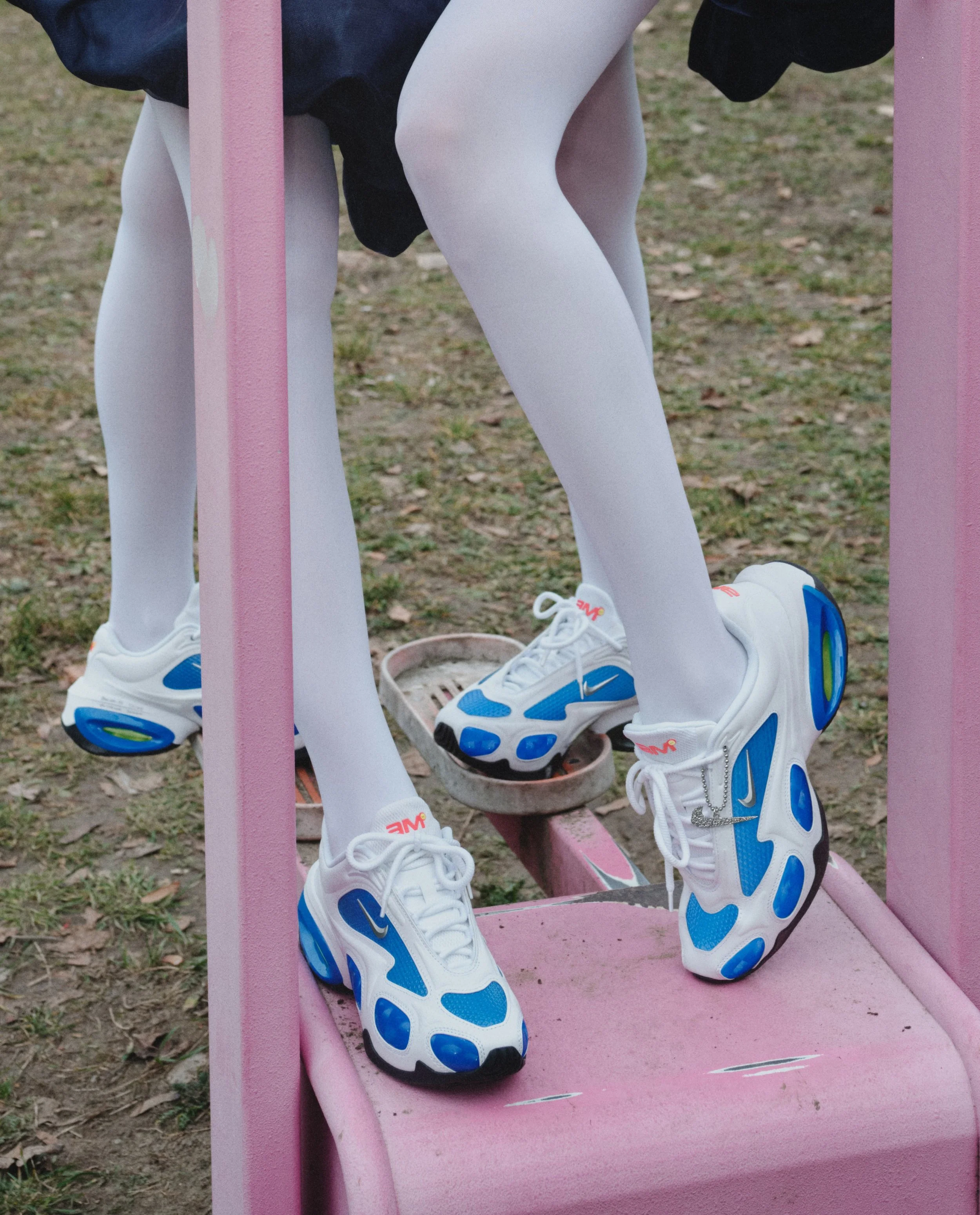 Close-up of a person wearing white leggings and white, blue, and black sneakers, sitting on a pink park bench with grass and soil ground visible.