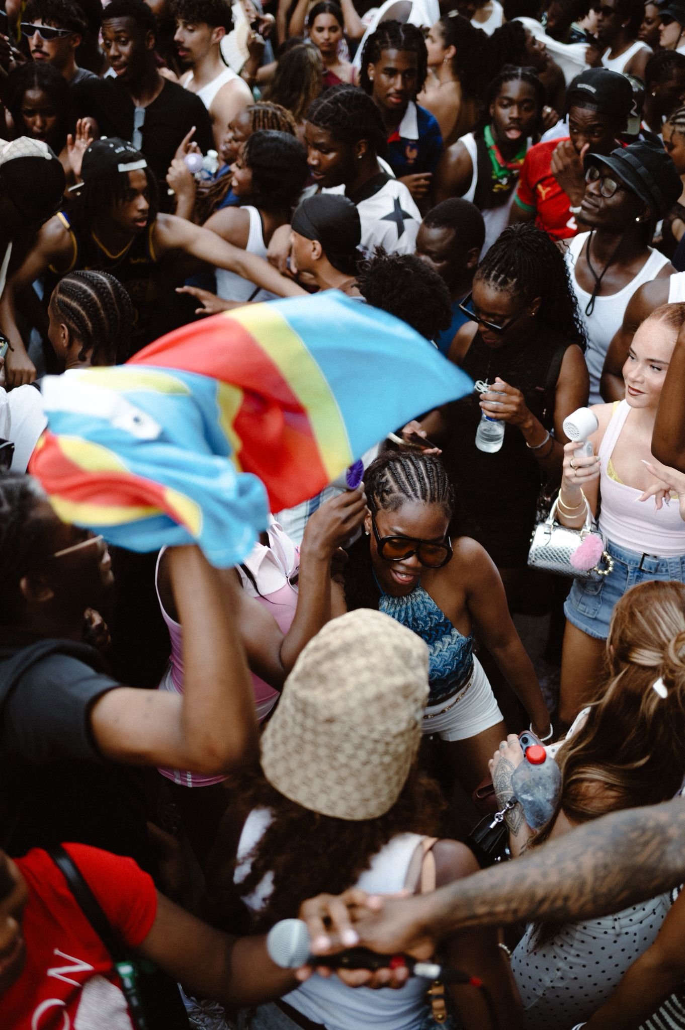 A large crowd of diverse people gathered closely together at an outdoor event. Some are holding drinks, flags, and personal items. Many appear to be engaged, taking photos or talking.