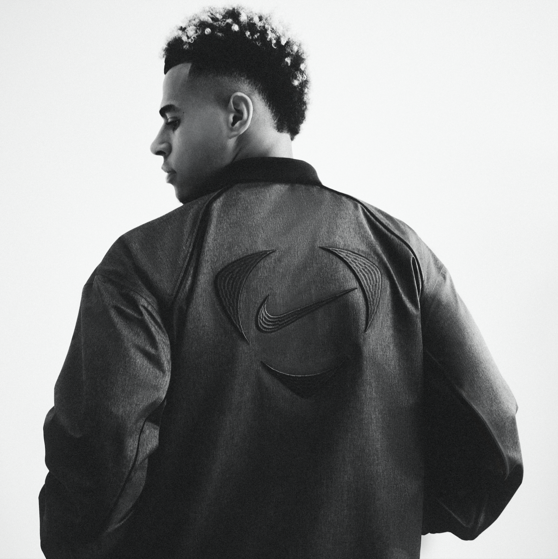 A black-and-white photo of a young man in profile, wearing a leather jacket with a Nike logo on the back.