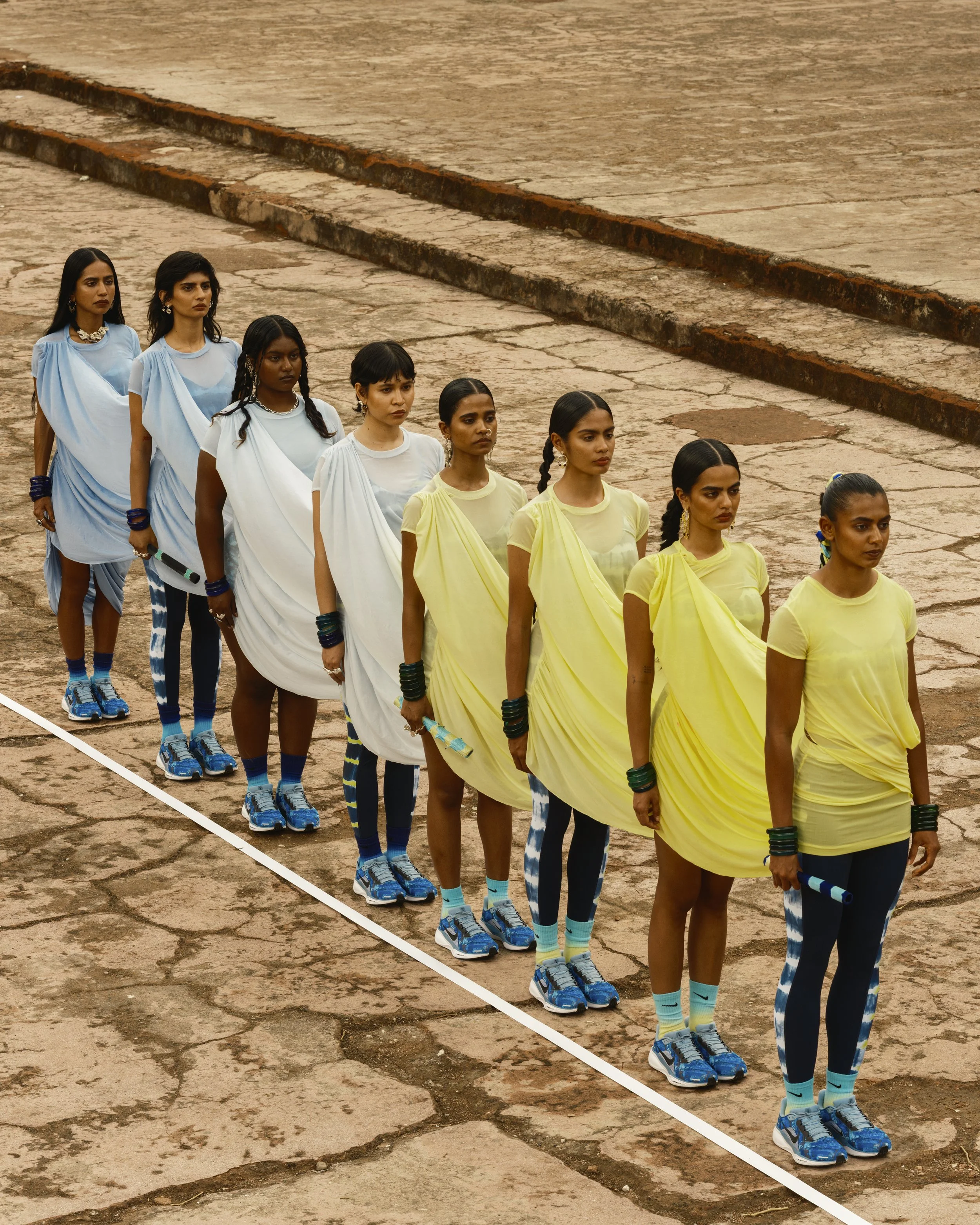 A line of nine women dressed in robes and athletic shoes, holding batons, standing on a cracked stone surface.