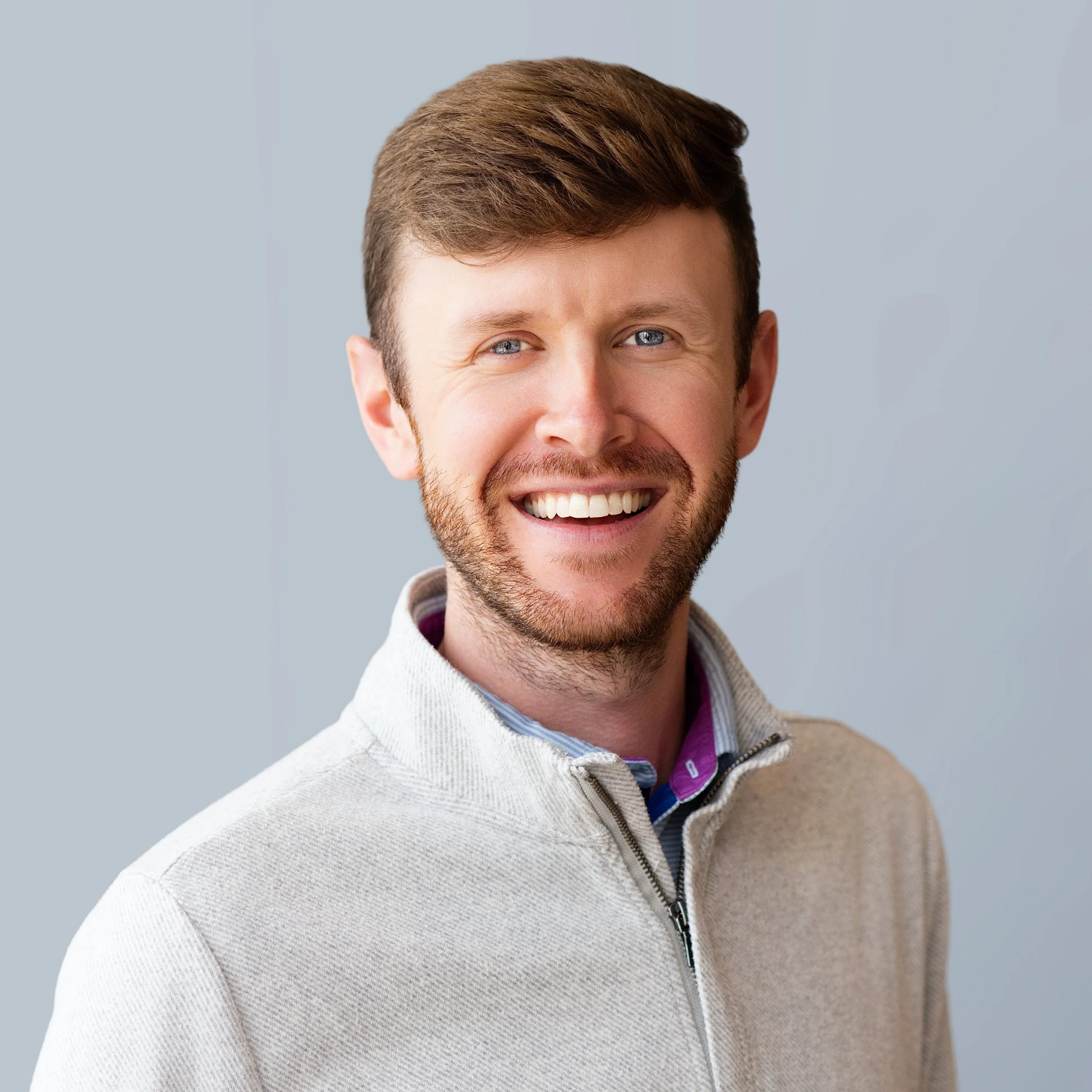 Close-up of a smiling man with brown hair, blue eyes, and a light beard, wearing a light-colored sweater with a zipper and a collared shirt underneath, against a plain gray background.