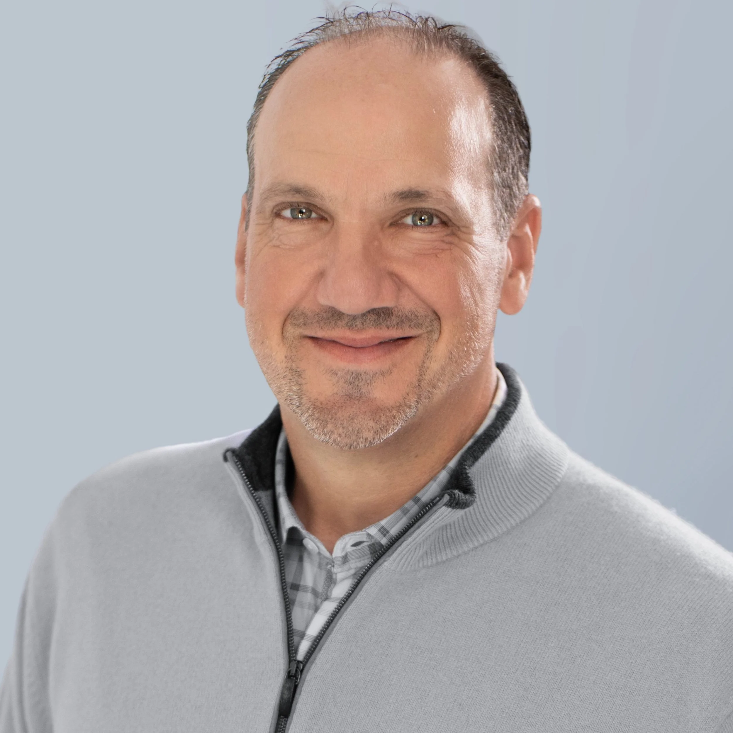 Close-up of a middle-aged man's face with short hair and a beard, smiling, wearing a gray zip-up sweater over a collared shirt, against a light gray background.