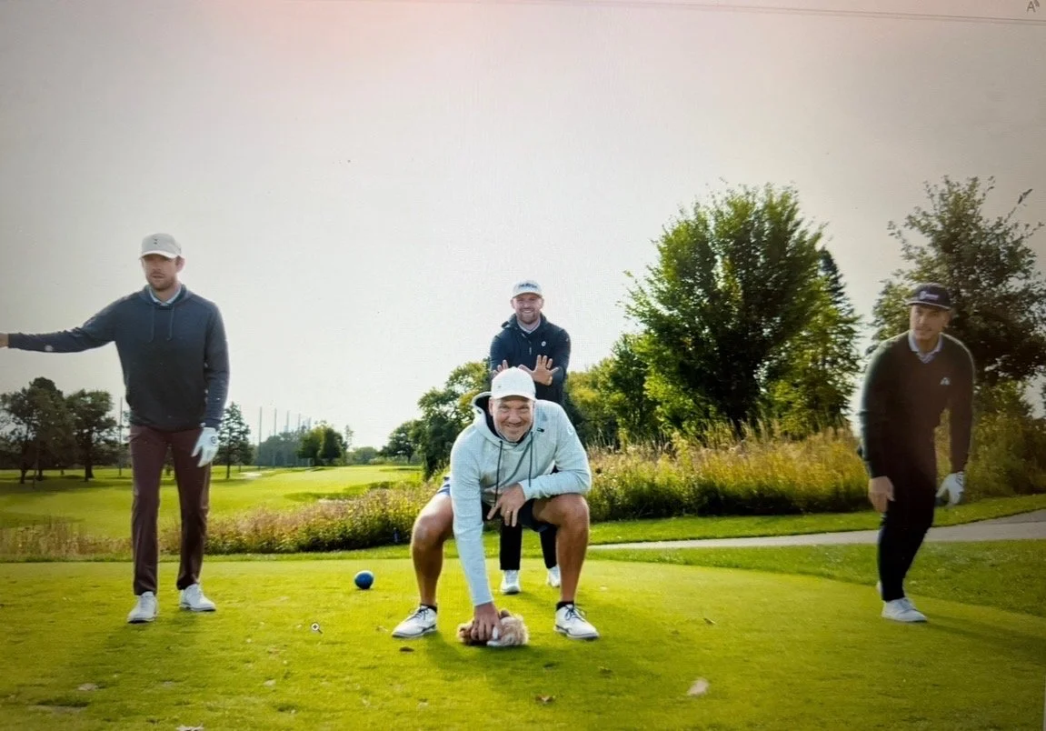 Four men on a golf course posing for a photo, with trees and a golf green in the background.