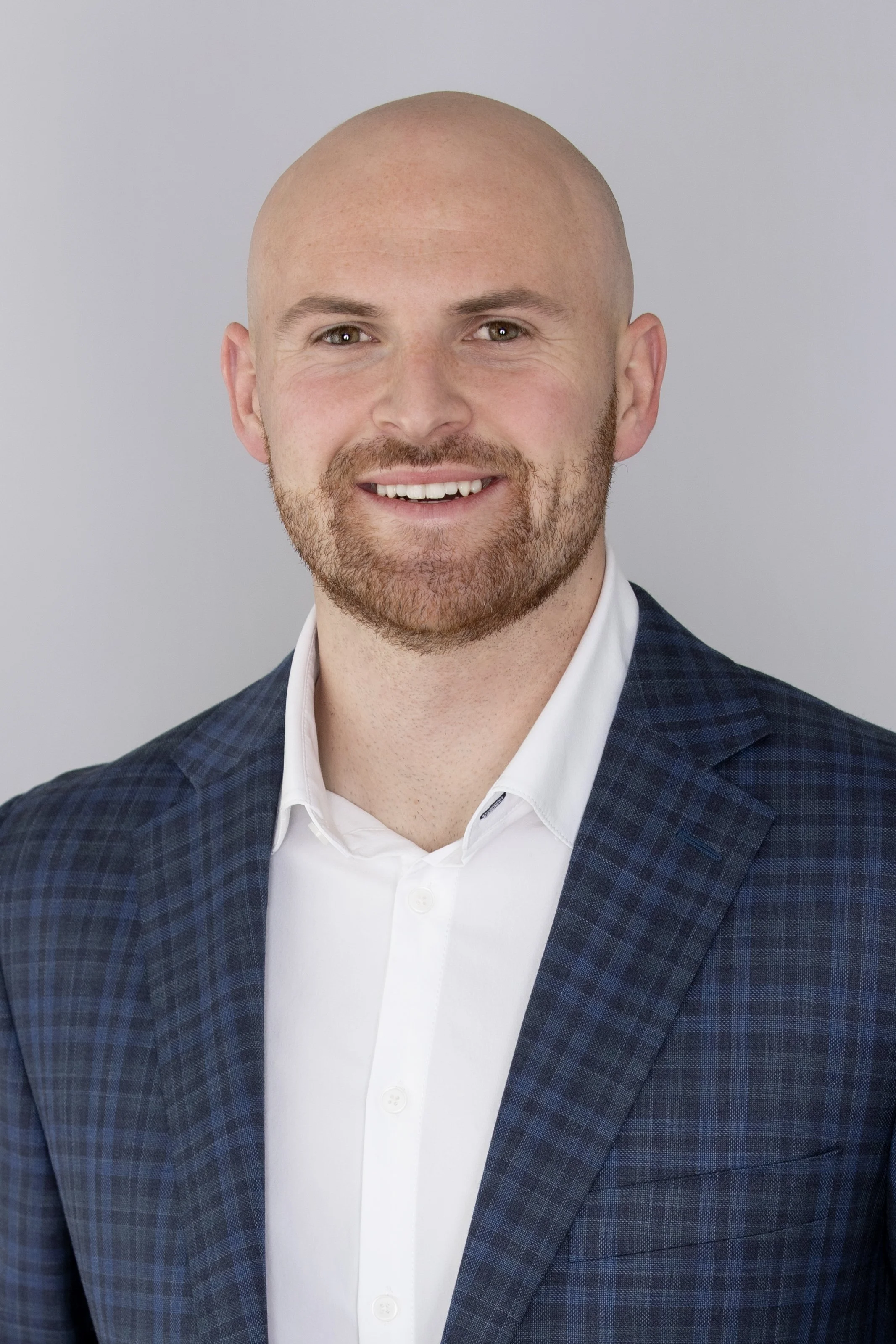 A smiling man with a beard and shaved head, wearing a white shirt and a blue checkered blazer against a light gray background.