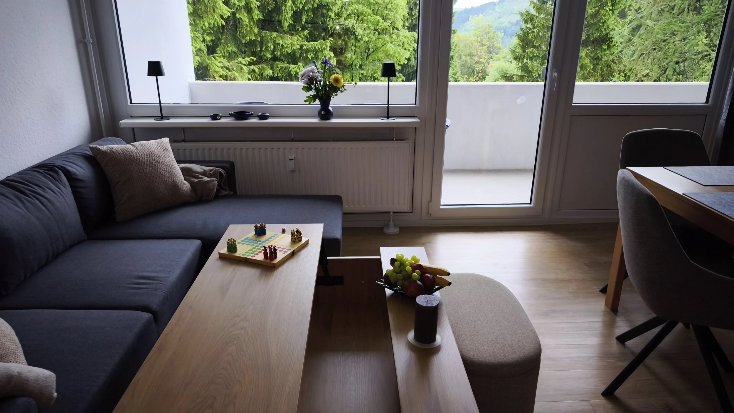 Wohnzimmer mit einem grauen Sofa, einem runden Tisch mit Obst und einem Brettspiel, großen Fenstern, die eine grüne Landschaft zeigen, sowie einer Pflanzenvase auf der Fensterbank.