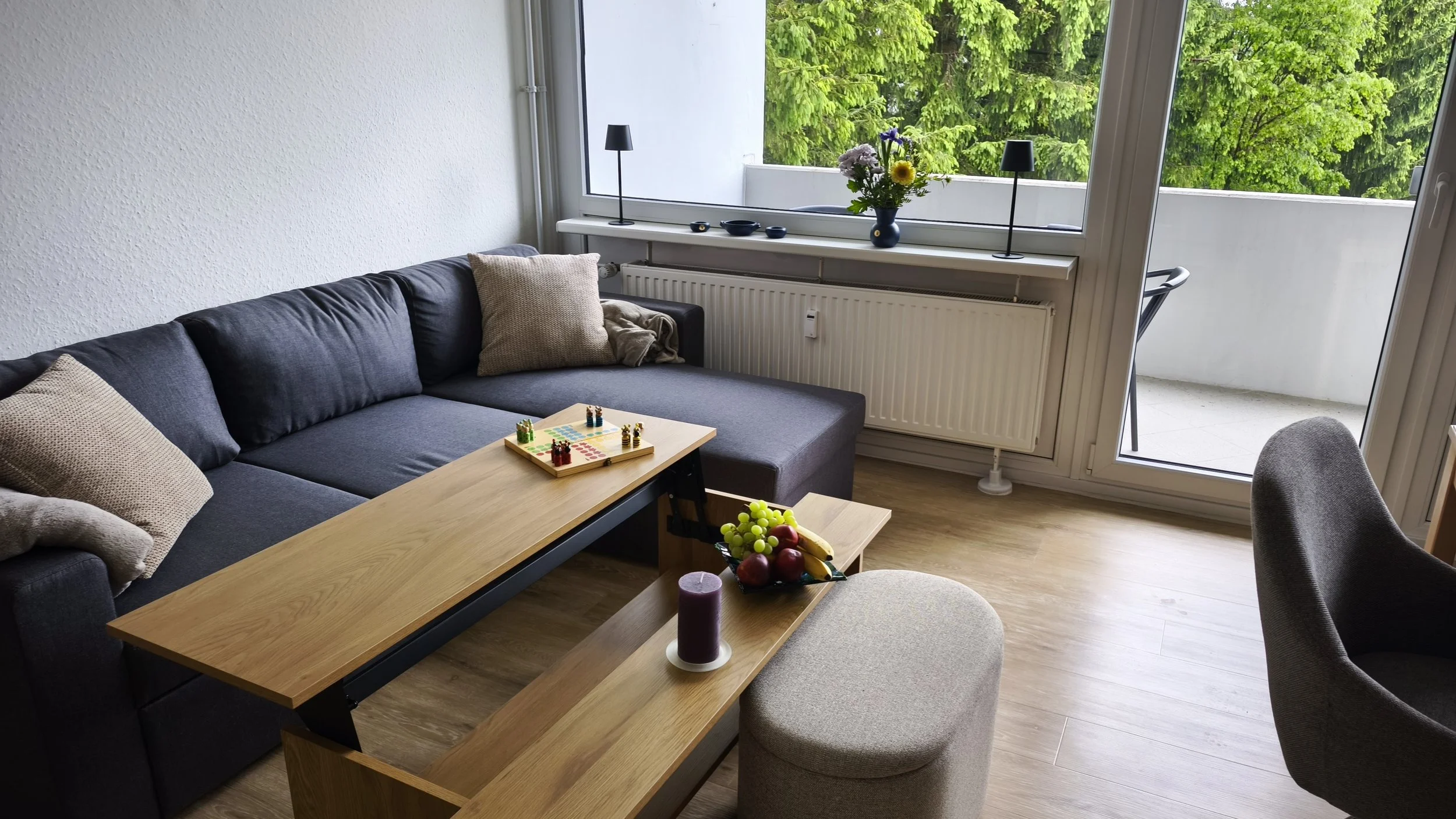 Wohnzimmer mit grauem Sofa, Kissen, Couchtisch mit Spiel, Obst und Kerze, Fenster mit grünen Bäumen im Hintergrund.