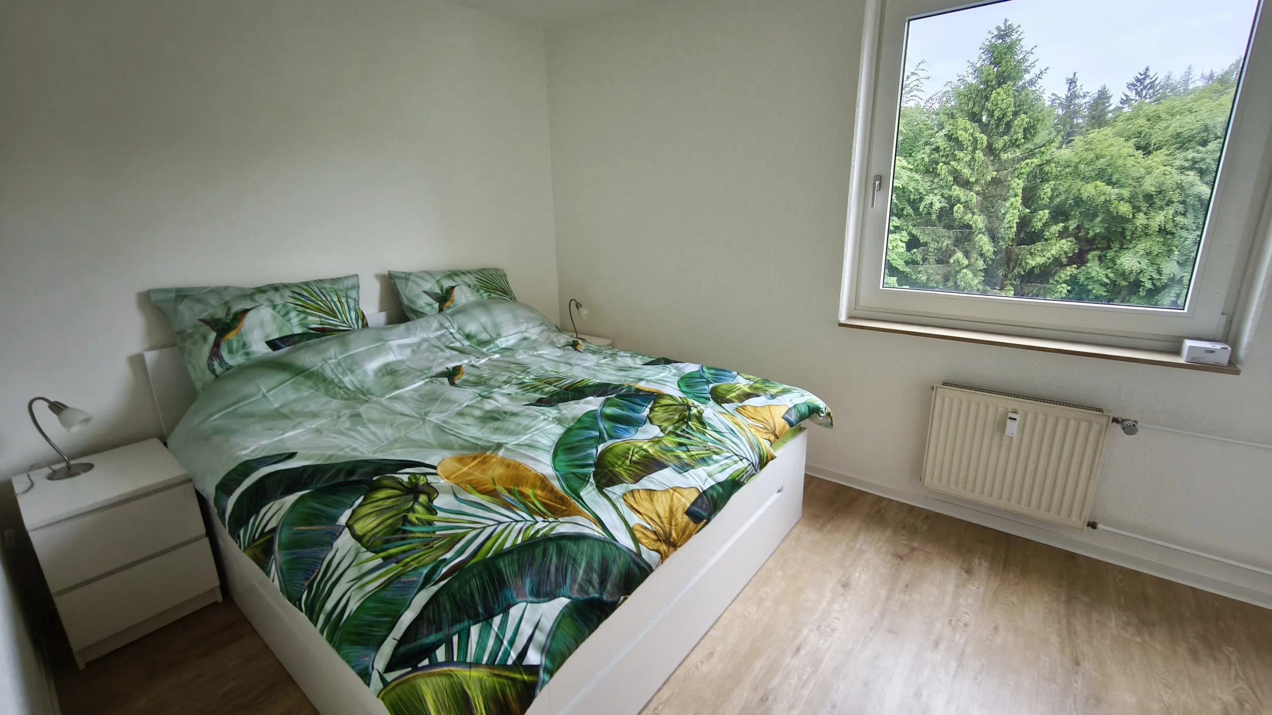 Ein Schlafzimmer mit einem weißen Bett, das mit einem grünen Bettwäsche-Set mit Blatt- und Vogelmotiven bedeckt ist. Es gibt eine weiße Nachttischlampe auf einer weißen Nachttischkommode und ein großes Fenster mit Blick auf grüne Bäume. An der Wand unter dem Fenster befindet sich eine Heizung.