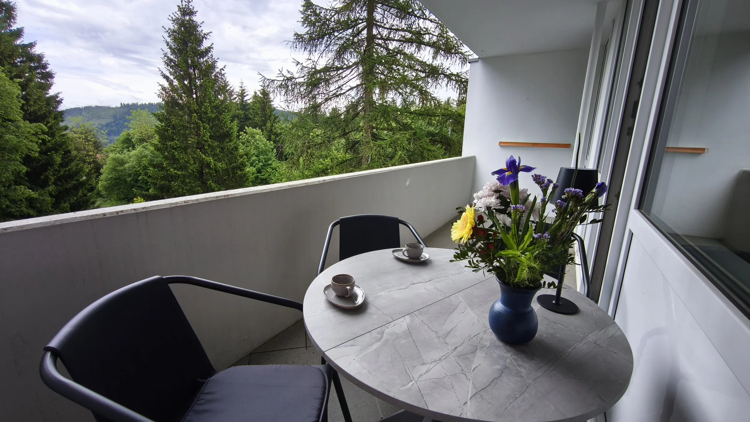 Balkon mit einem runden, weißen Tisch, einem blauen Blumenstrauß, drei Tassen und Stühlen, mit Blick auf grüne Bäume und Hügel unter einem bewölkten Himmel.
