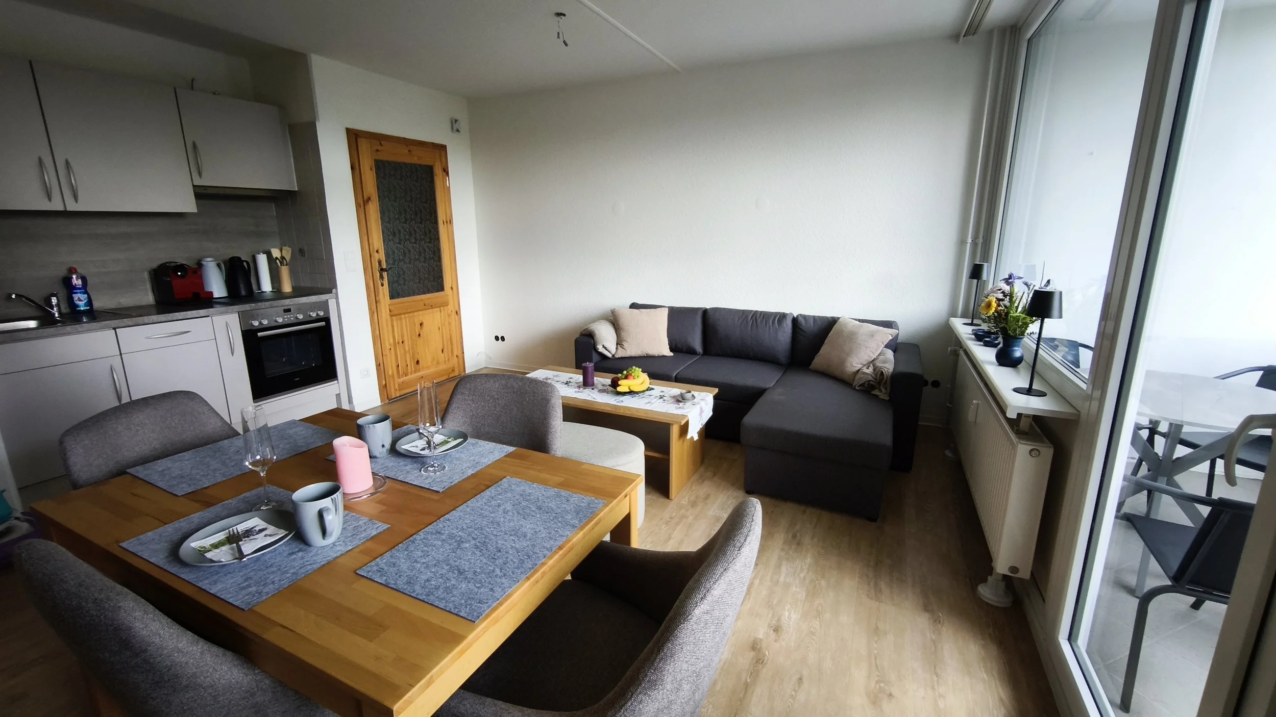 Offener Wohn- und Essbereich mit Küche, Esstisch, Couch, Couchtisch, Fenster mit Blumen, Balkon mit Terrasse im Hintergrund