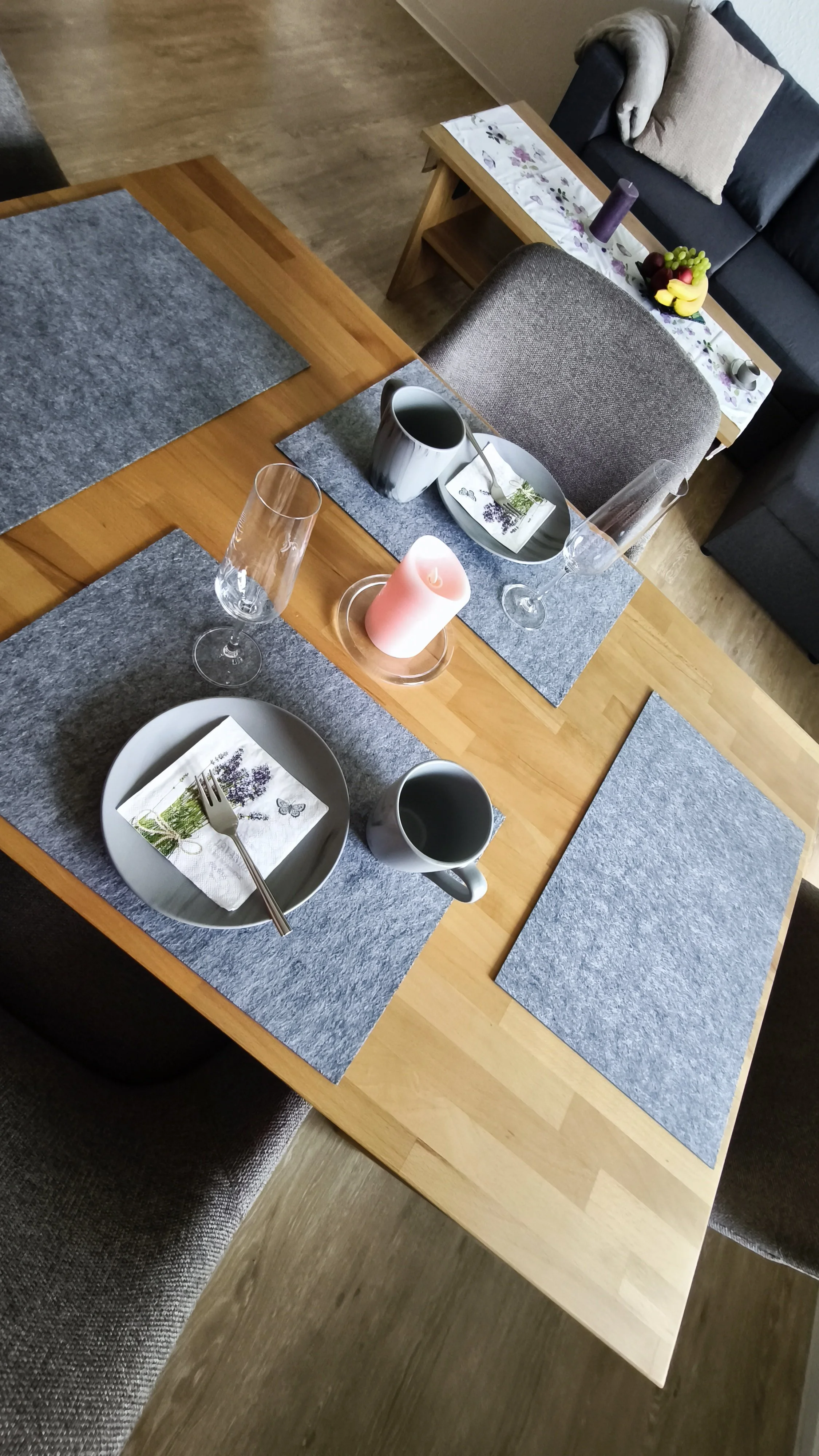 Ein gedeckter Esstisch mit grauen TABLETTEN, weißen Tellern, Silberbesteck, schwarzen und weißen Tassen, Weingläsern und Kerzen, im Hintergrund festlicher Tisch mit Obst und Kerzen auf der Couch.
