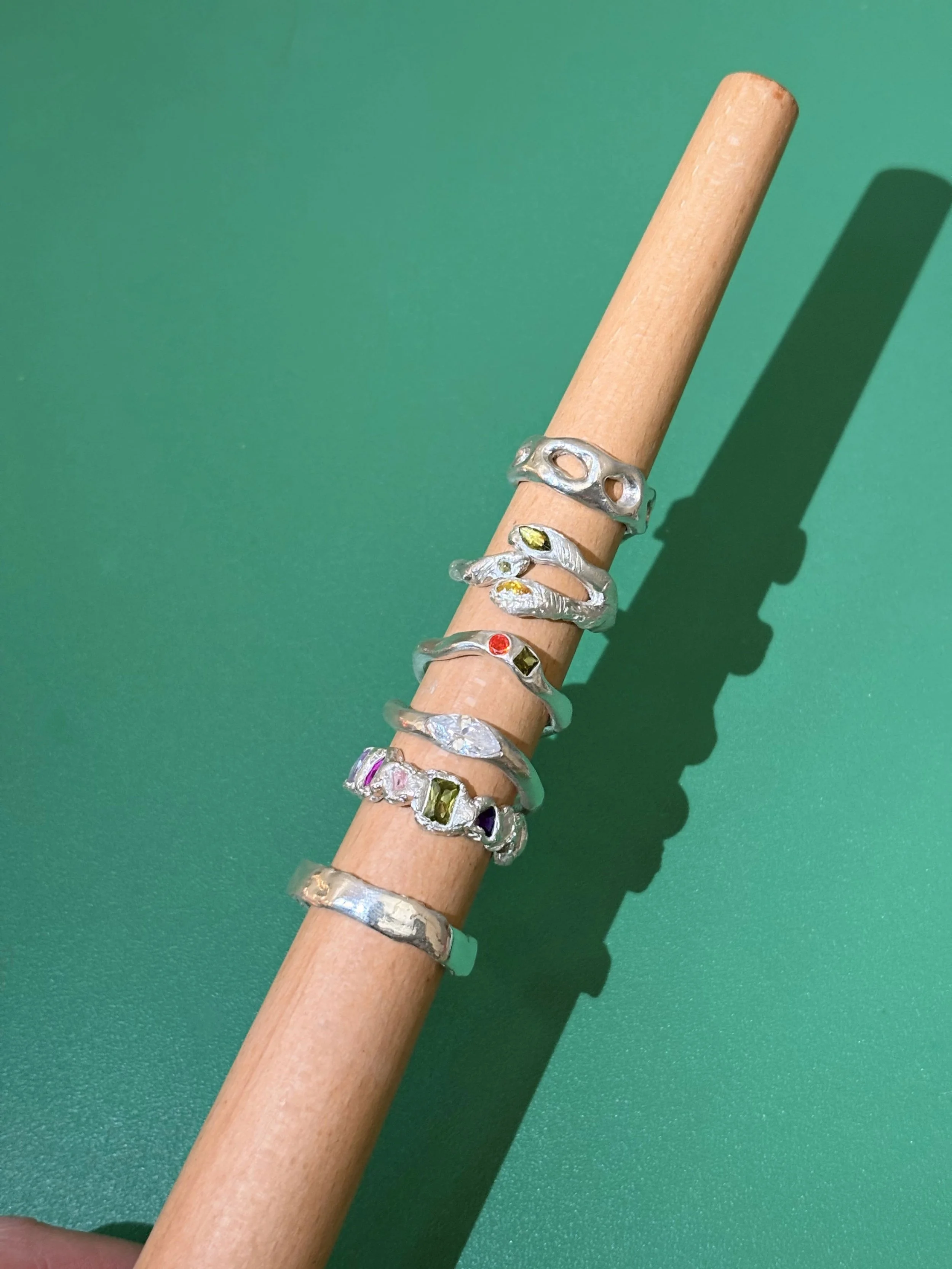 Une main tient un bâton en bois avec plusieurs bagues en argent ornées de pierres colorées, le tout sur un fond vert.
