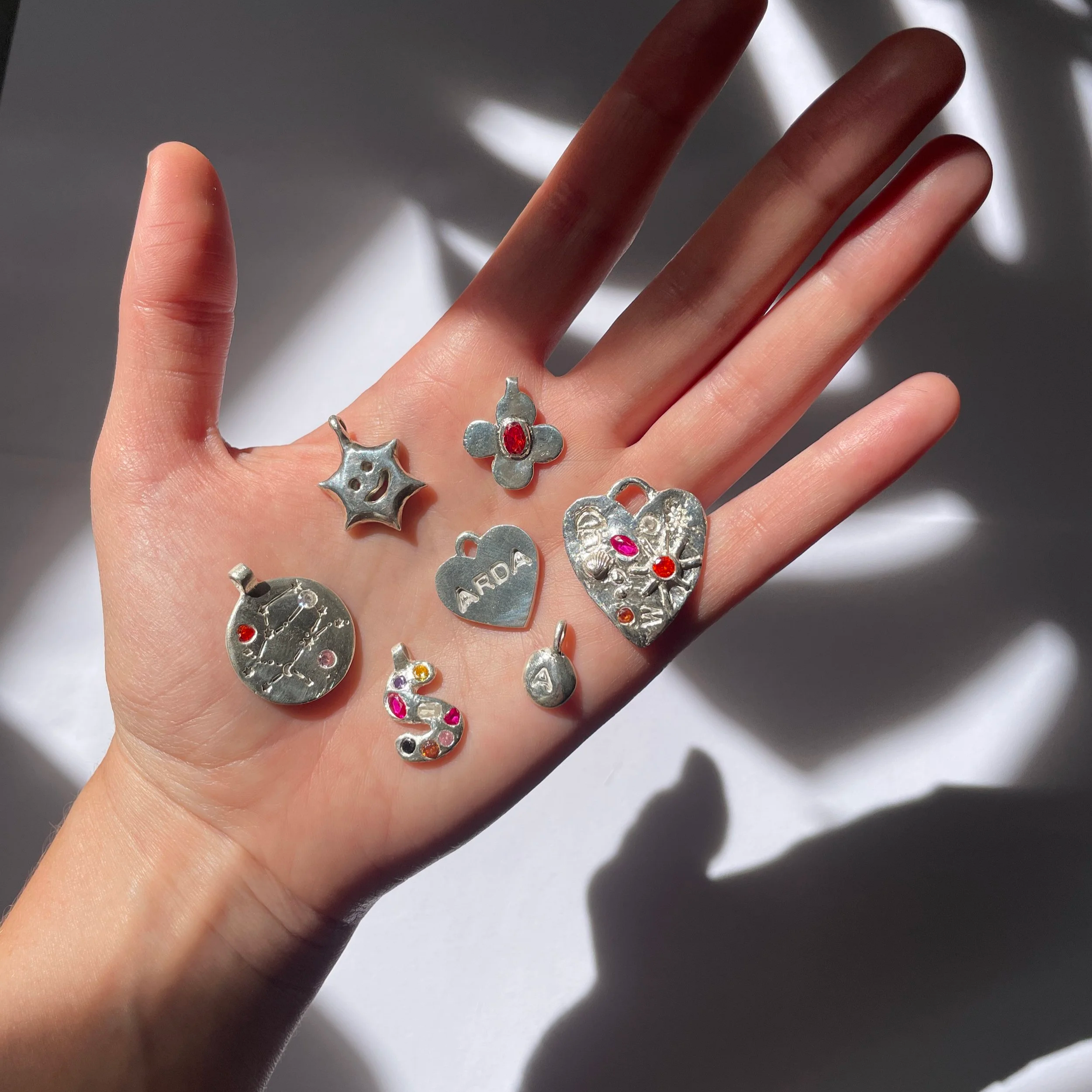 Main d'avec plusieurs bagues en argent, dont une avec une pierre rouge, et des dessins de constellations et de petits cœurs, avec ombres de lumière sur un fond gris.