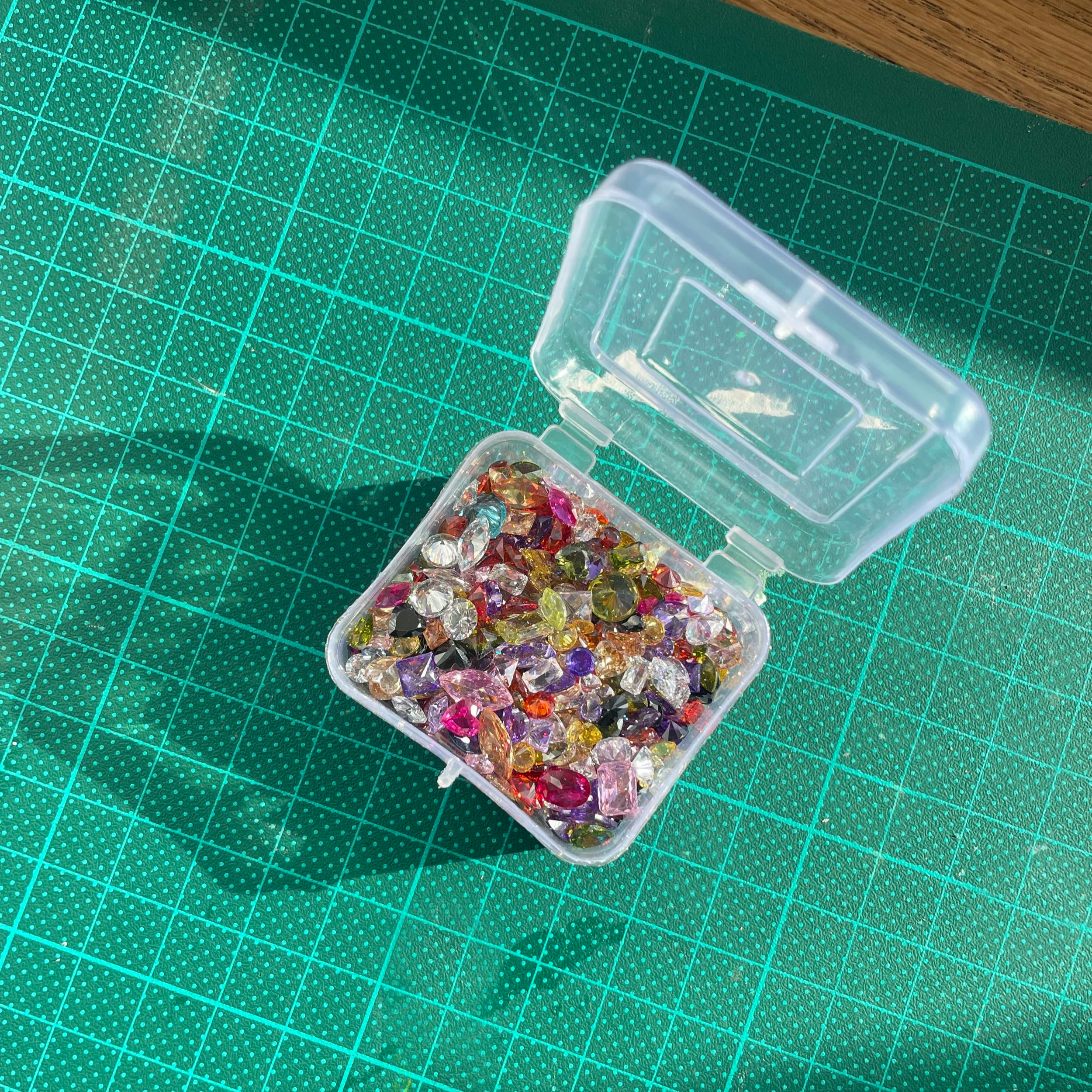 Une boîte en plastique transparente contenant des pierres précieuses colorées, posée sur une planche de découpe verte avec un motif de points