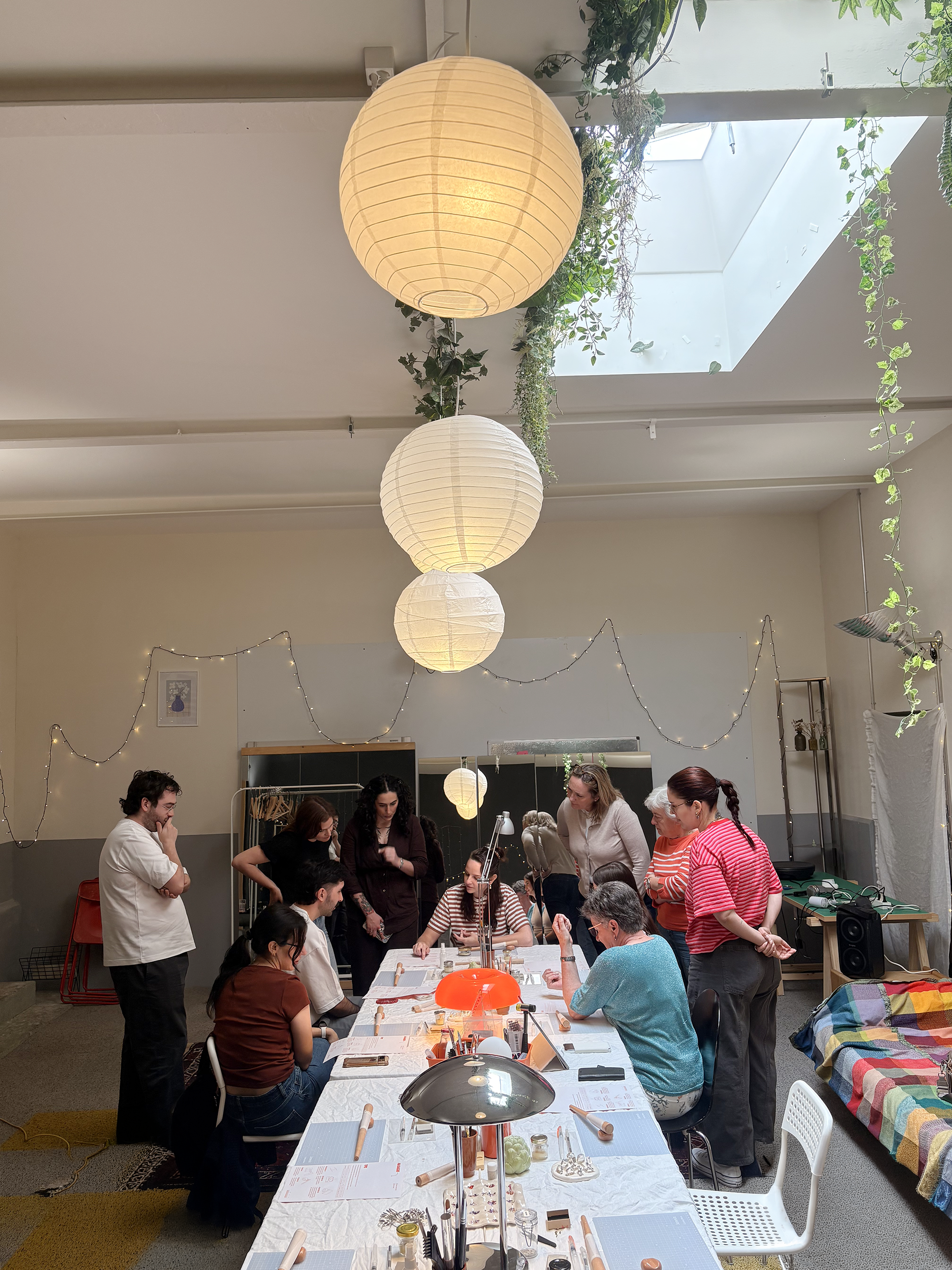Groupe de personnes autour d'une table, probablement lors d'un atelier ou d'une classe d'artisanat, dans une salle bien éclairée avec des lampes en papier et des plantes en décoration.