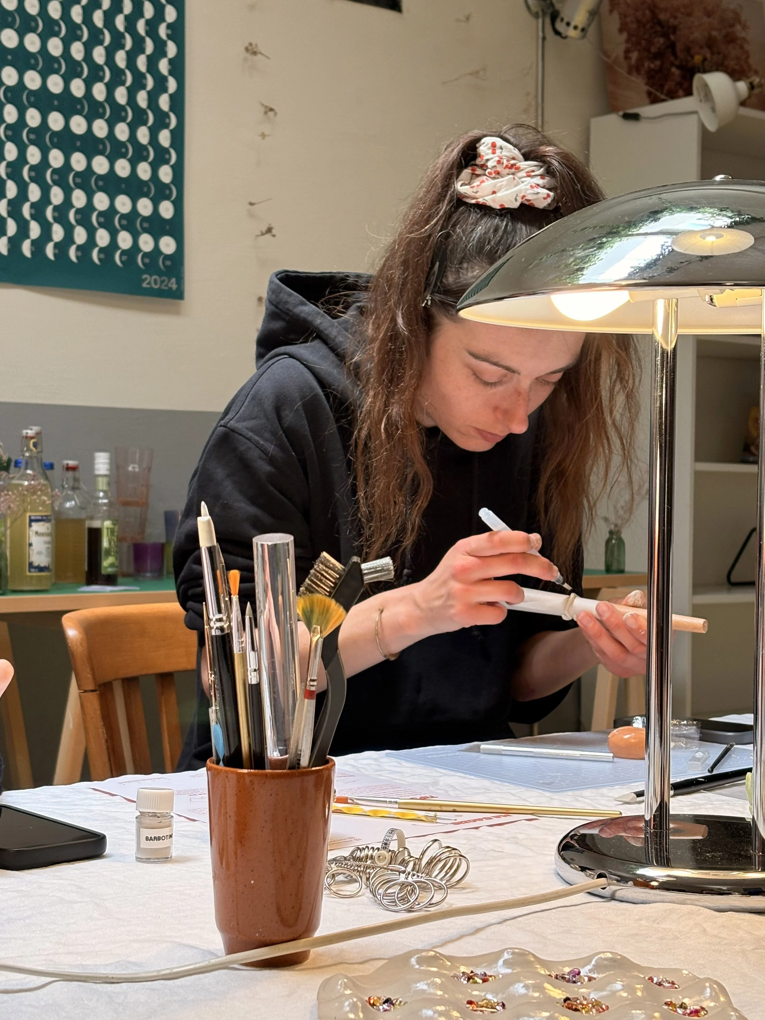 Une femme réalise une création artistique ou artisanale sur un pied, assise à une table de travail, avec divers outils et matériaux autour d'elle, dans un atelier ou un espace de création.