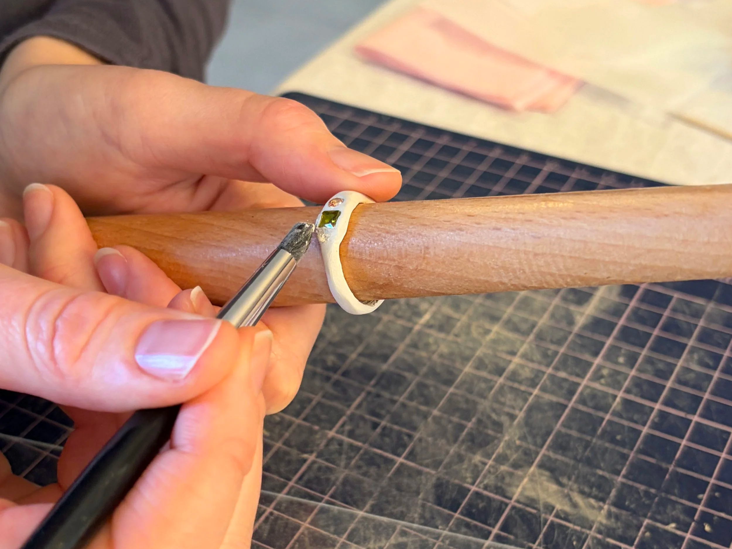 Une personne utilise un pinceau pour déposer un accessoire avec une pierre précieuse sur une bague en en argent, posée sur une surface en bois.