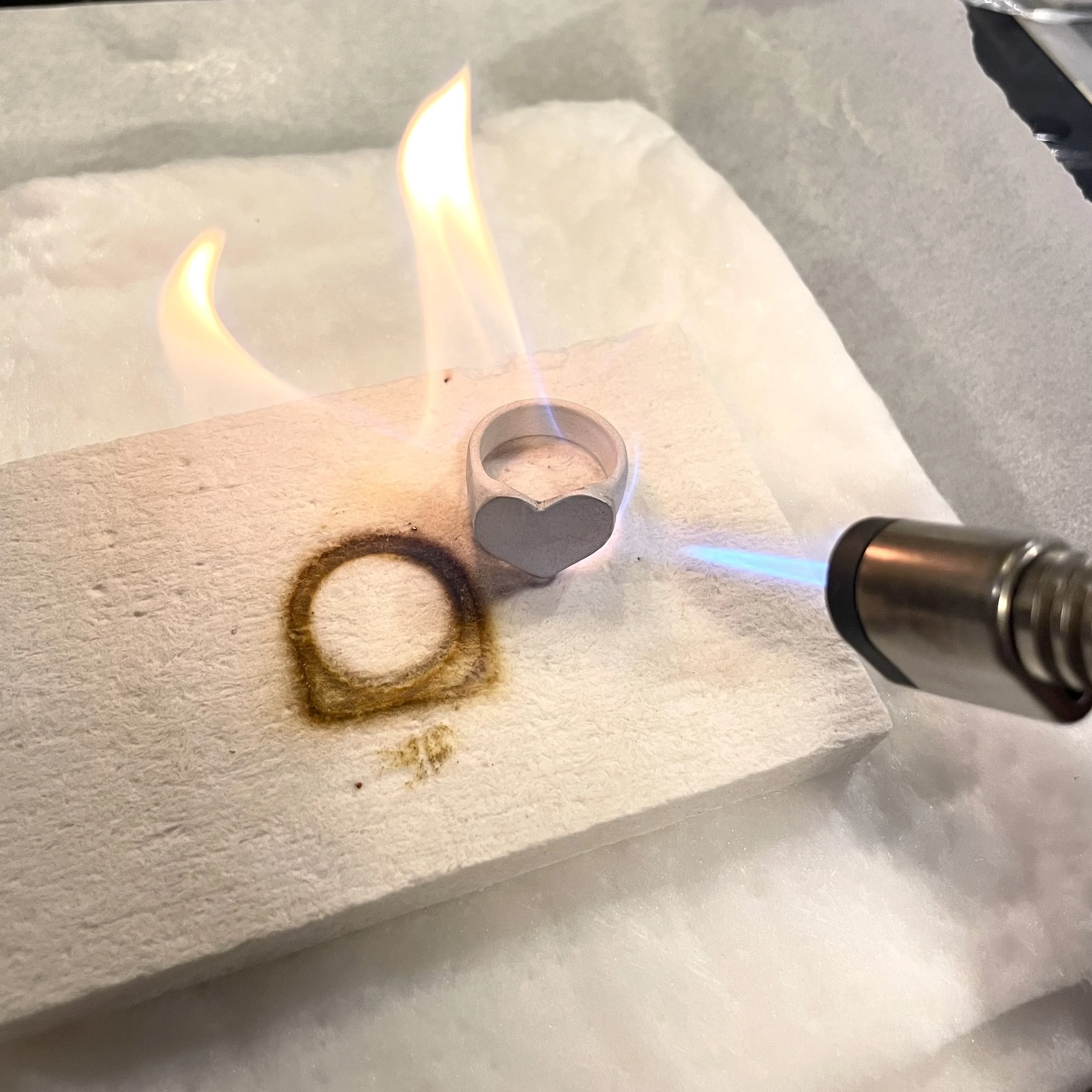 Une flamme de brûleur à gaz sur une surface blanche, une pièce en métal est visible à côté.