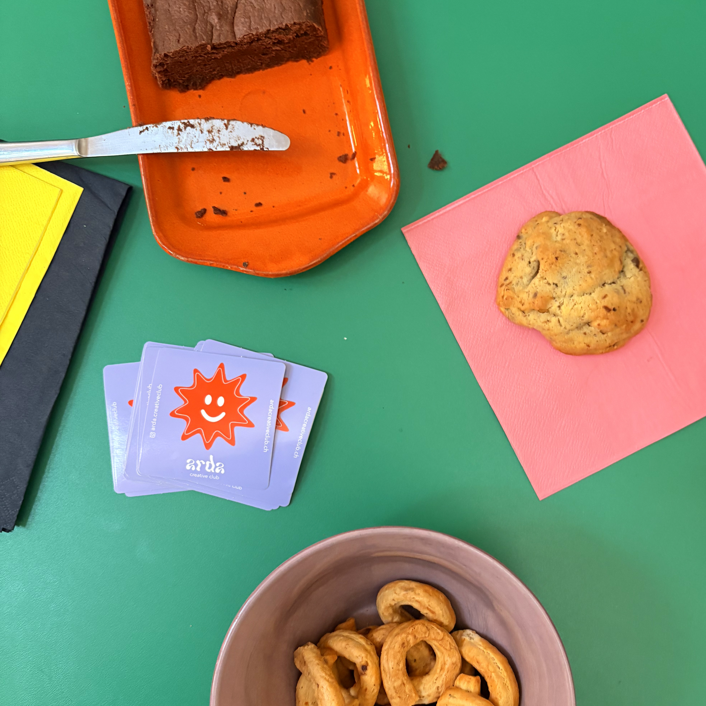 Une table verte avec un bol en céramique contenant des biscuits, une assiette orange avec un gâteau au chocolat et une petite pile de cartes de visites d'Arda Creative Club. Sur la table, il y a aussi des serviettes en papier rose.